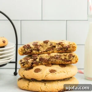 A stack of beautifully thick gluten-free Crumbl chocolate chip cookies, one broken open to reveal its gooey, melted chocolate center. A glass of milk with a striped straw sits nearby, completing the irresistible dessert scene.