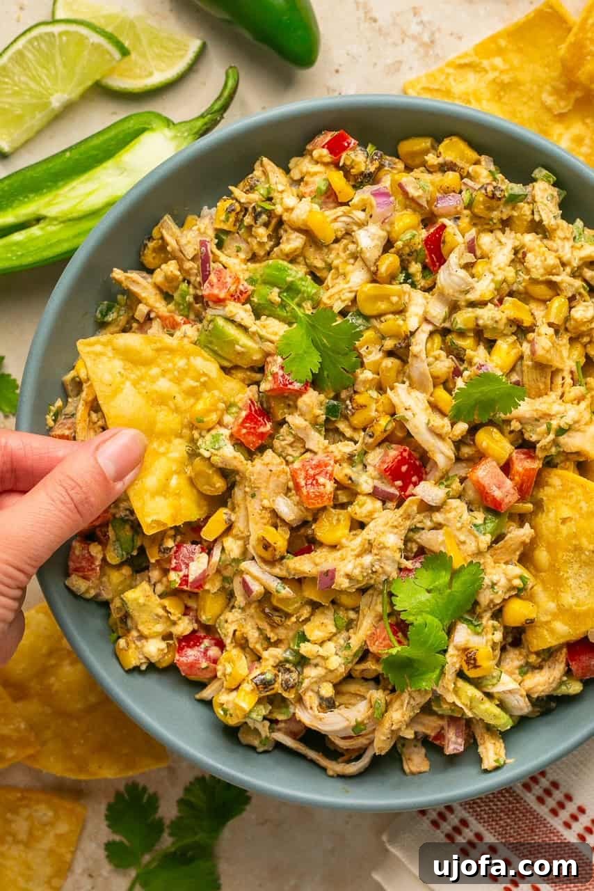 A bowl of Mexican chicken salad with a hand dipping a chip into it, showcasing the vibrant ingredients.