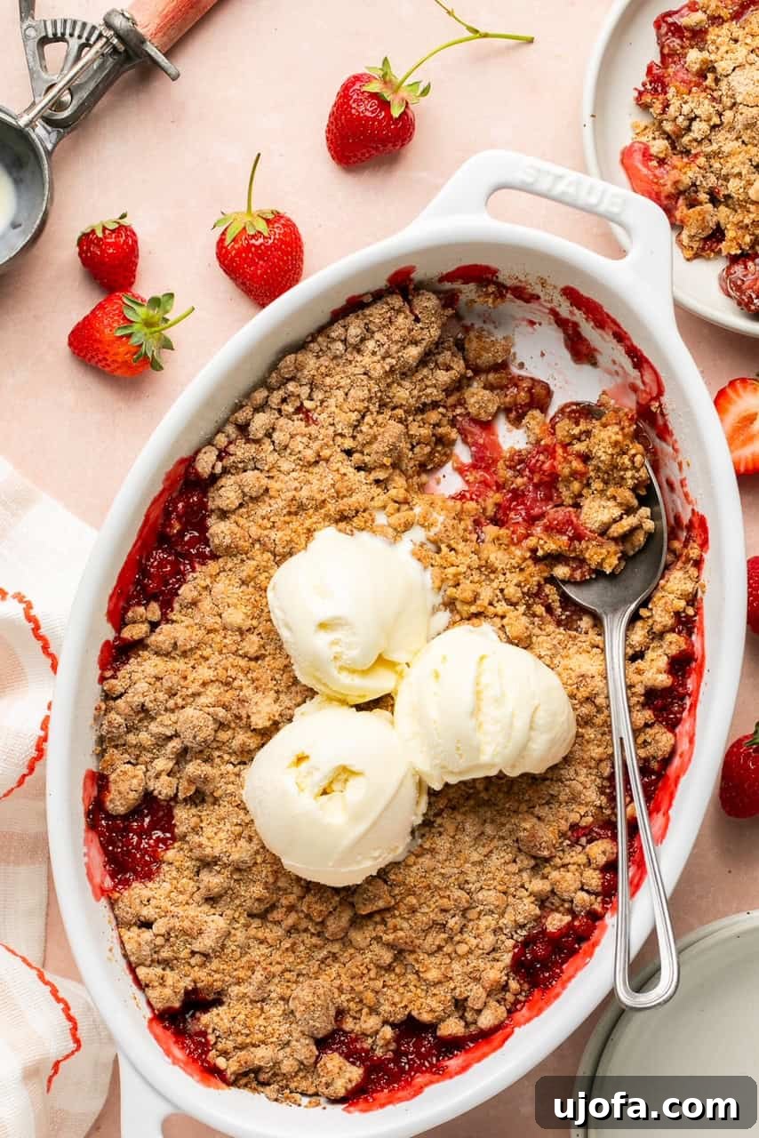 Sweet & Simple Gluten-Free Strawberry Crumble 2 A beautifully baked strawberry crumble in an oval baking dish, generously topped with three scoops of creamy vanilla ice cream. A spoon is poised, ready to serve a portion of this irresistible summer dessert.