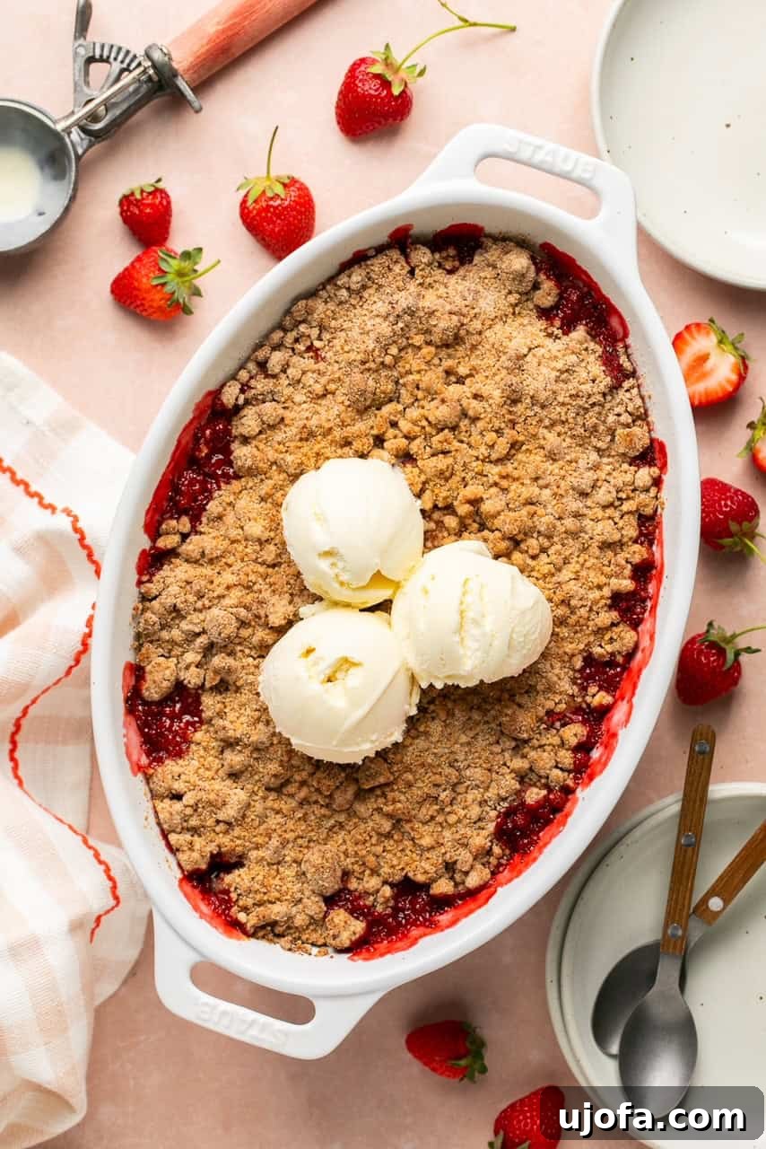 Sweet & Simple Gluten-Free Strawberry Crumble 7 An inviting overhead shot of a freshly baked strawberry crumble, presented in a white baking dish and adorned with three scoops of creamy vanilla ice cream, ready to be enjoyed.
