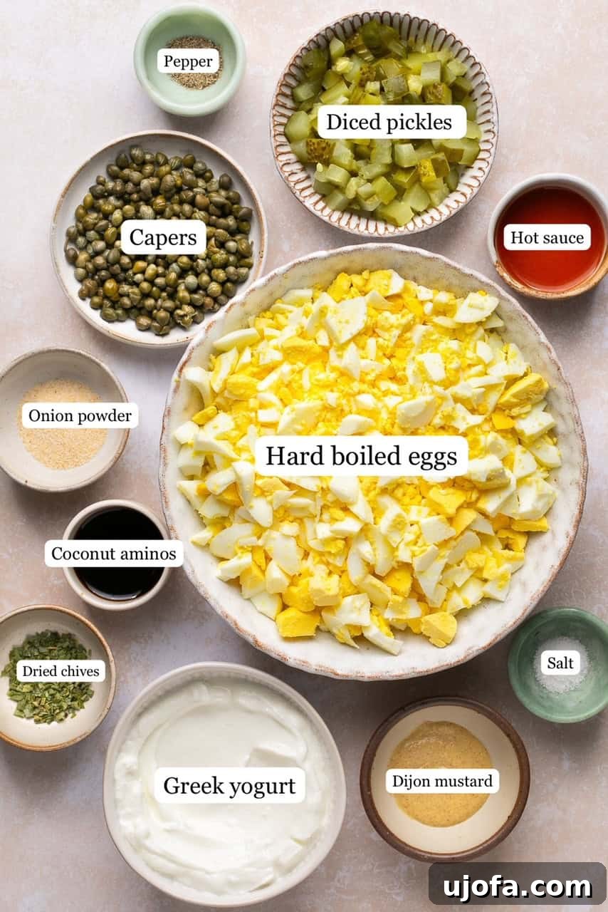 Recipe ingredients laid out in small, labeled bowls, showcasing fresh eggs, diced pickles, capers, yogurt, mustard, and seasonings.