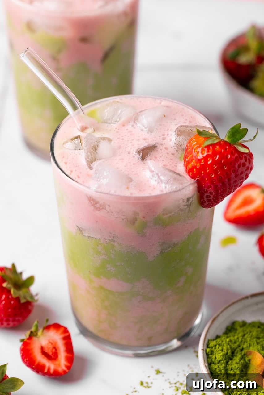 A glass of iced strawberry matcha tea latte, garnished with a fresh strawberry, showcasing its beautiful green and pink layers.