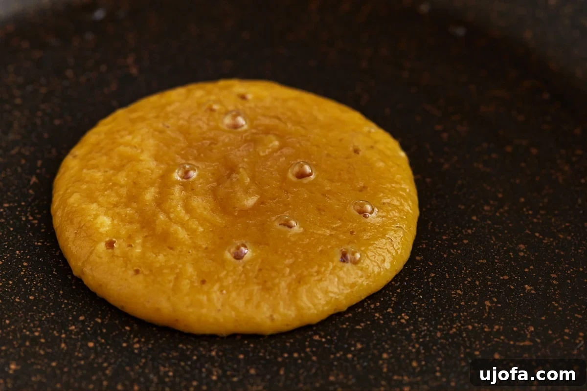 A coconut flour pancake cooking on a griddle, showing the edges setting and bubbles forming on the surface, indicating it's ready to flip.