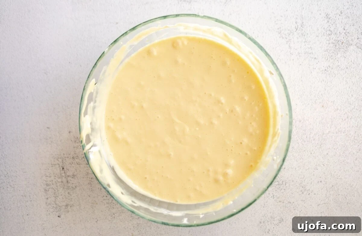 Silken Cheesecake 7 Fully mixed cheesecake batter with eggs incorporated, appearing smooth and pourable in a clear glass bowl, ready for the baking pan.