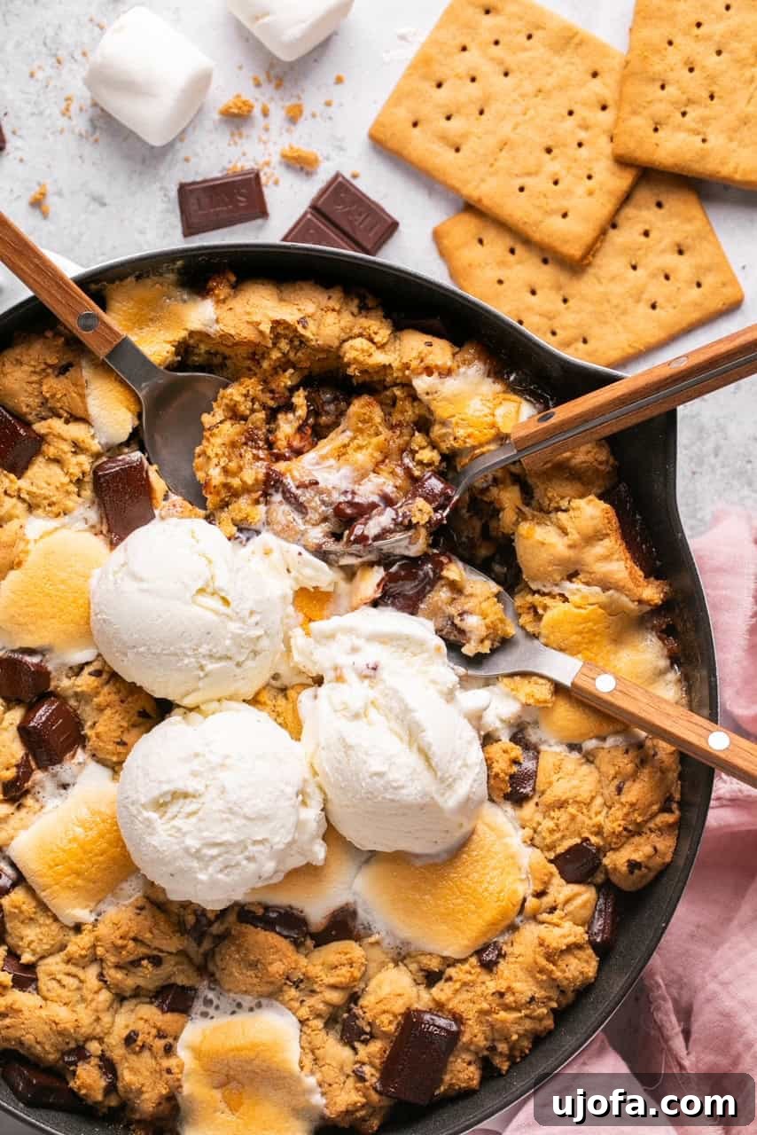 An overhead shot of a golden-brown chocolate and marshmallow cookie skillet in a cast iron pan, generously topped with melting vanilla ice cream and hints of graham cracker crumble.