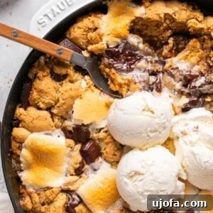 A s'mores chocolate and marshmallow cookie skillet topped with vanilla ice cream, with a spoon taking a bite.