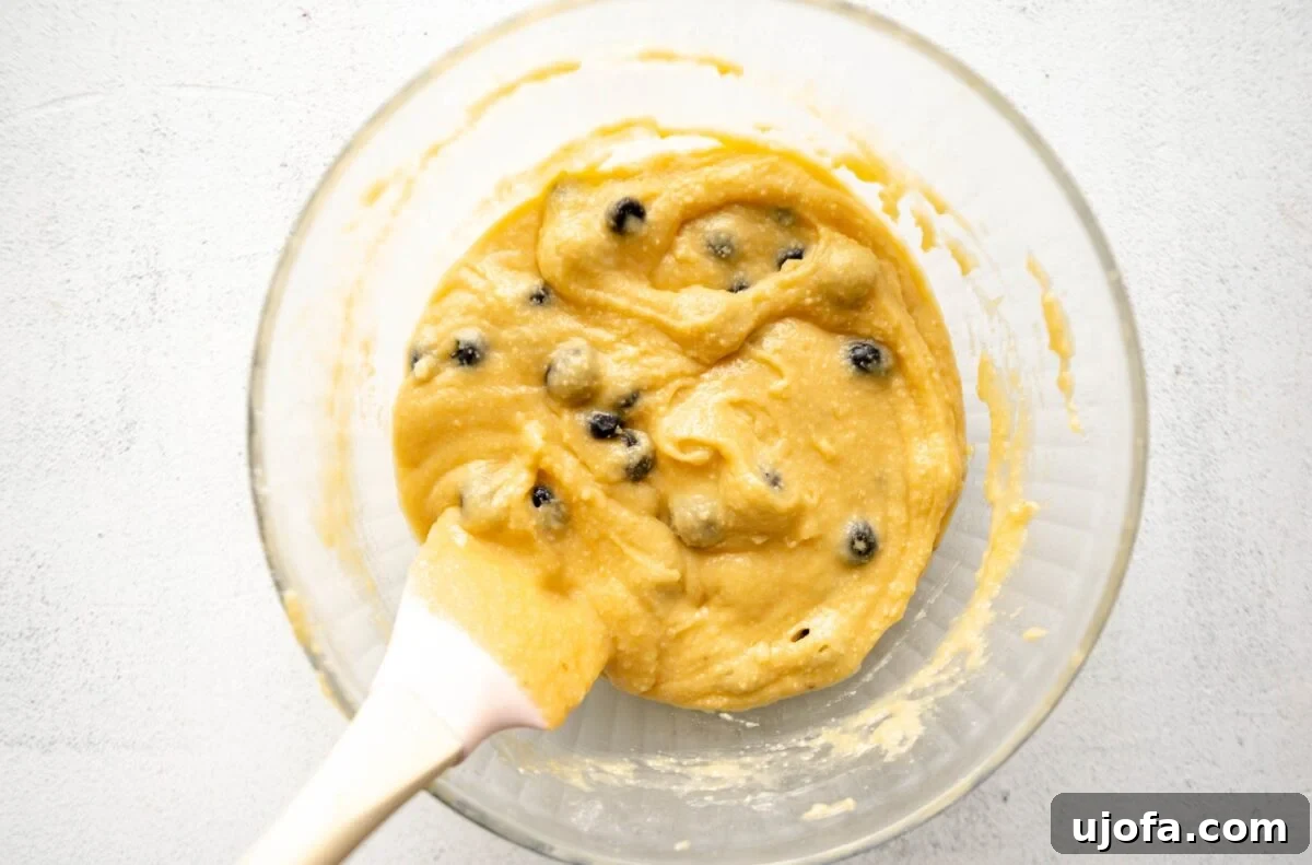 A clear glass bowl with almond flour muffin batter folded together with fresh blueberries, a white spatula resting inside the bowl.
