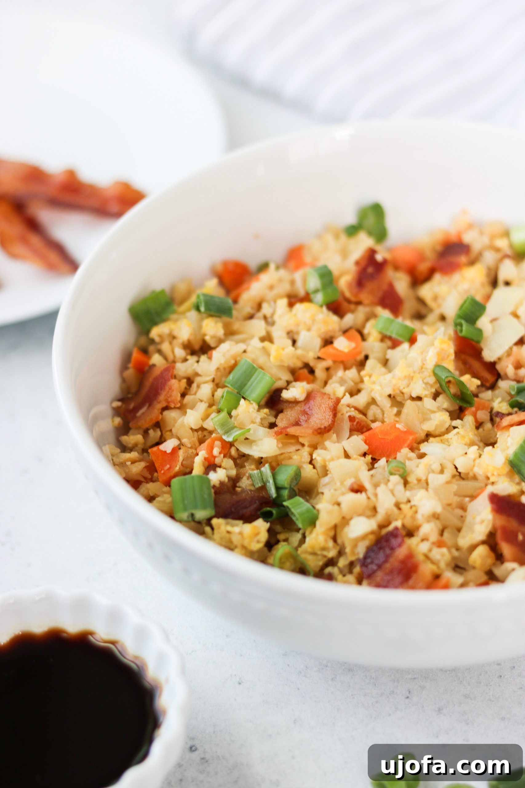 Close-up of a serving of Paleo Breakfast Fried Rice, highlighting the texture of cauliflower rice, bacon, and egg, garnished with fresh green onions.