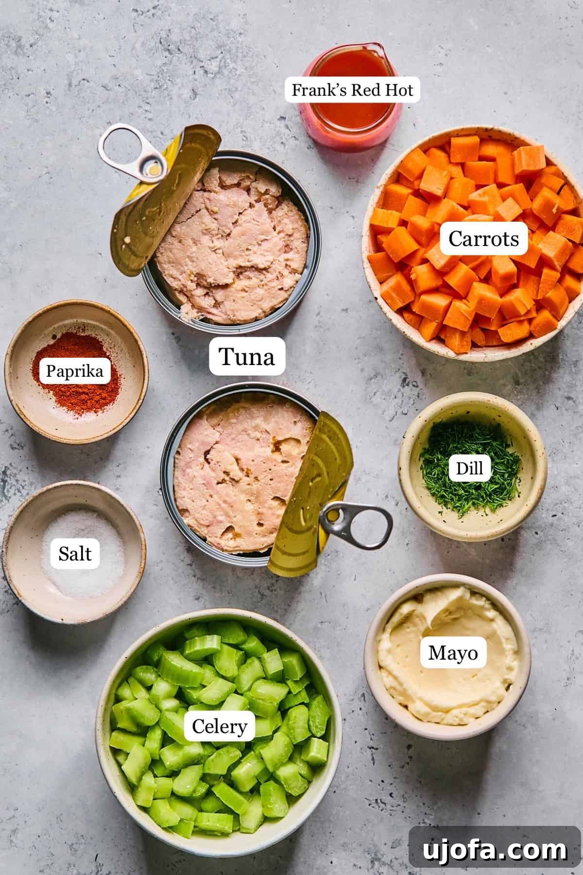 Small bowls containing labeled ingredients for buffalo tuna salad, including tuna, chopped celery, carrots, dill, and hot sauce