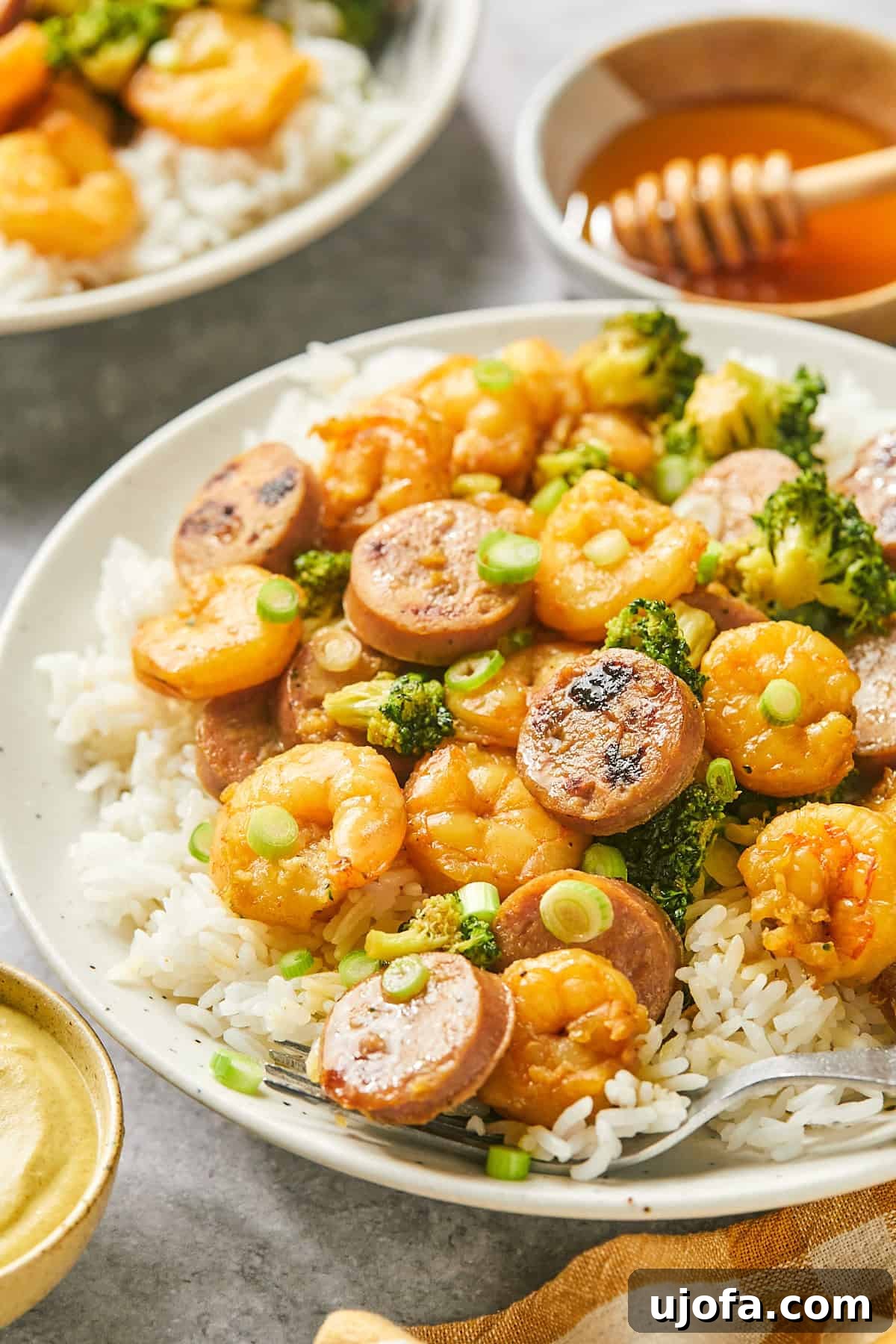 Plate of hot honey shrimp and sausage stir fry over white rice, with a small bowl of hot honey and green onions in the background.