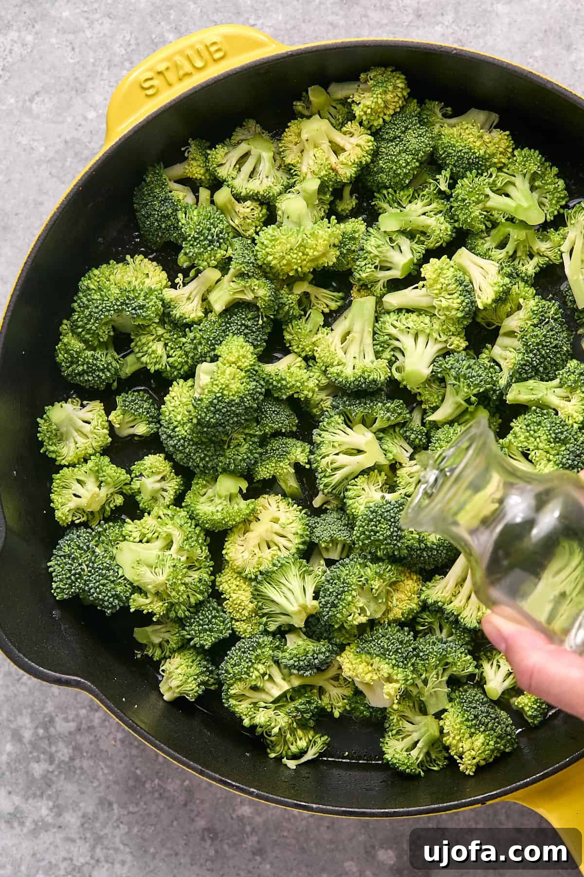Fresh broccoli florets in a skillet, with water being poured over them for steaming.