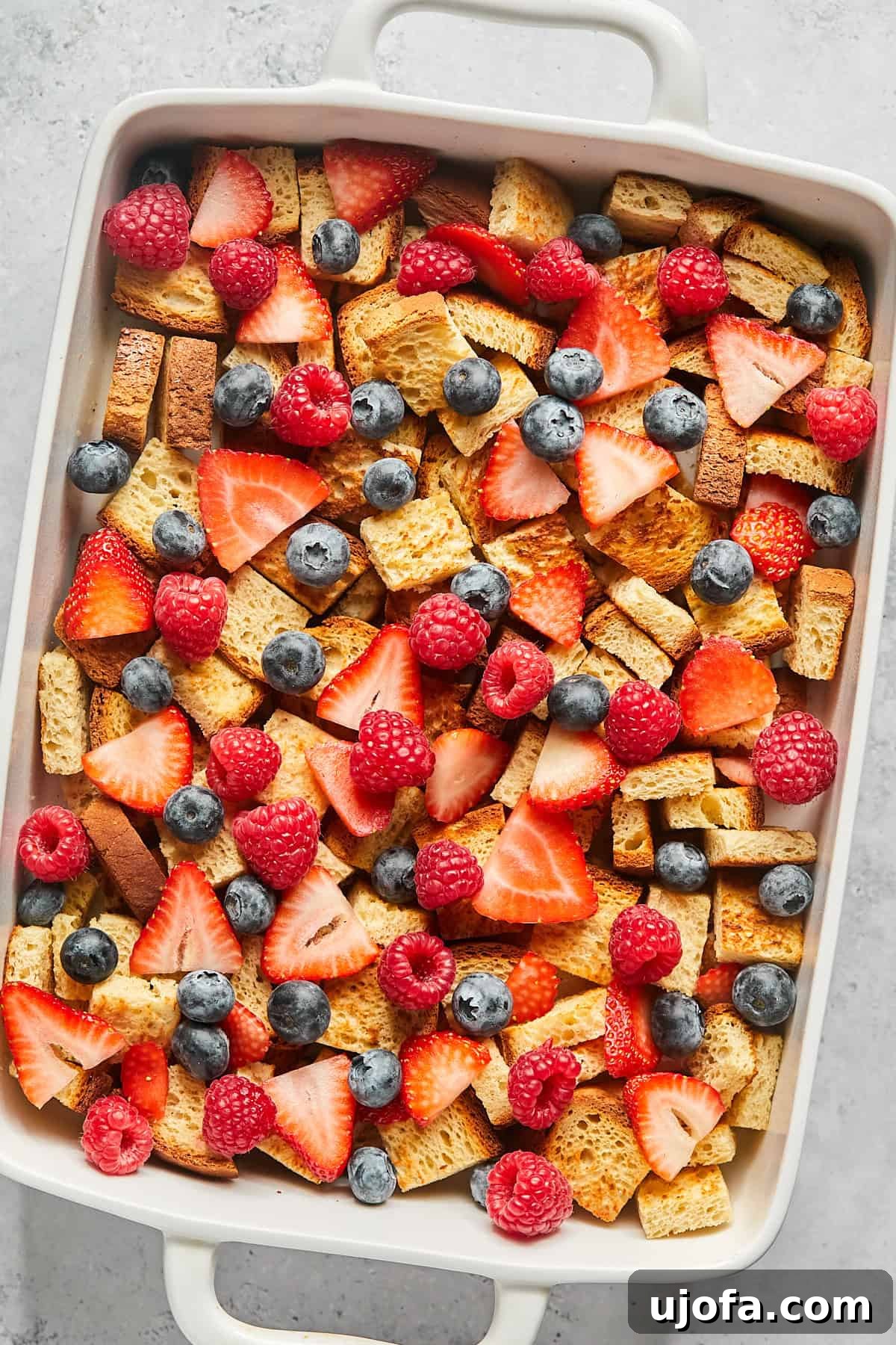 A colorful layer of fresh berries sprinkled over the toasted bread cubes in a casserole dish.