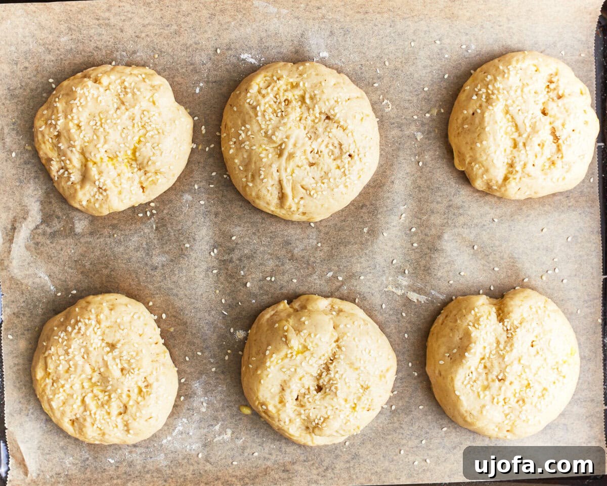 Burger Bliss Without The Gluten 10 Risen gluten-free hamburger bun dough on a baking sheet, topped with sesame seeds and ready for baking.