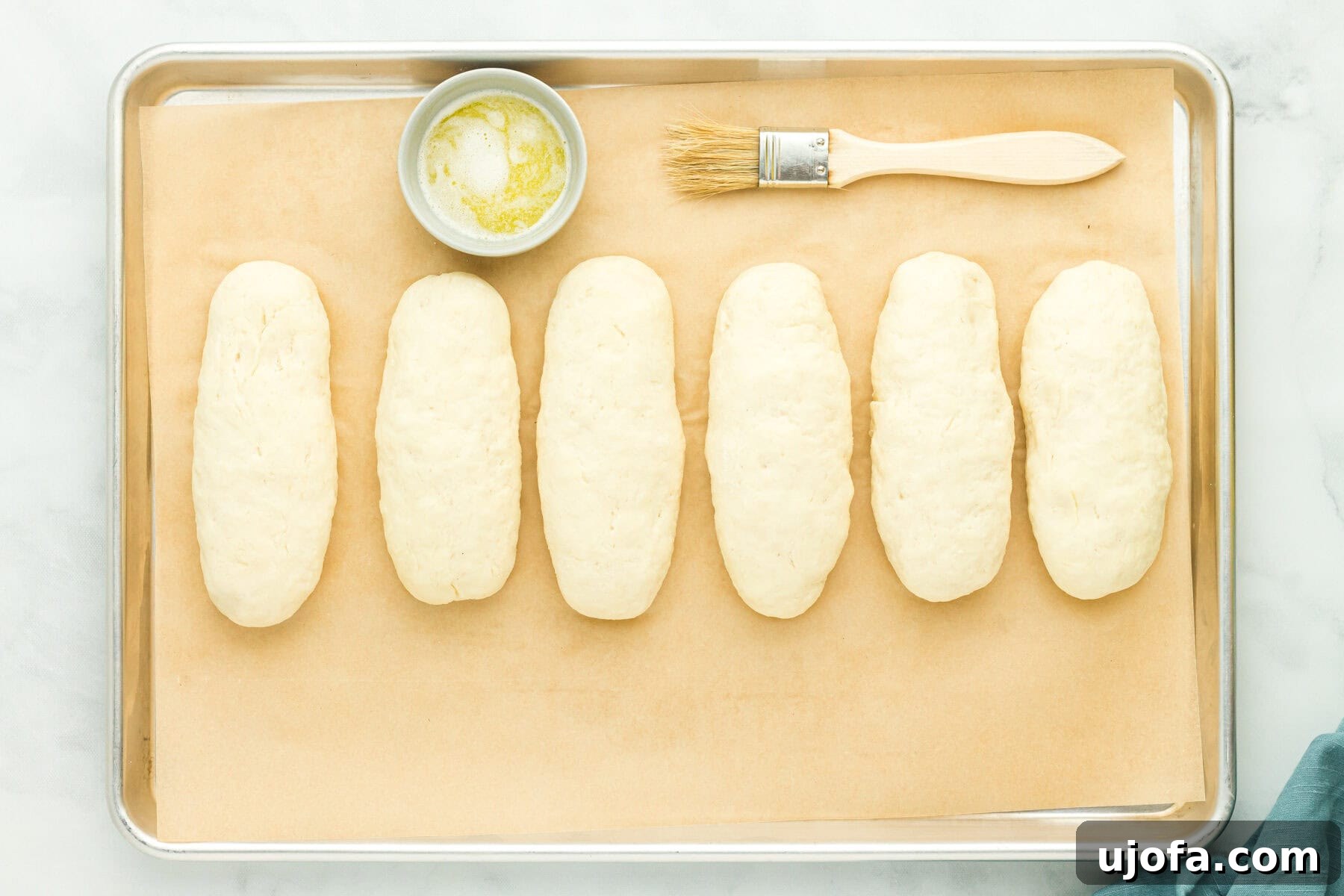 Gluten-free hot dog bun dough shaped into logs on a parchment lined baking sheet, alongside melted butter and a basting brush.