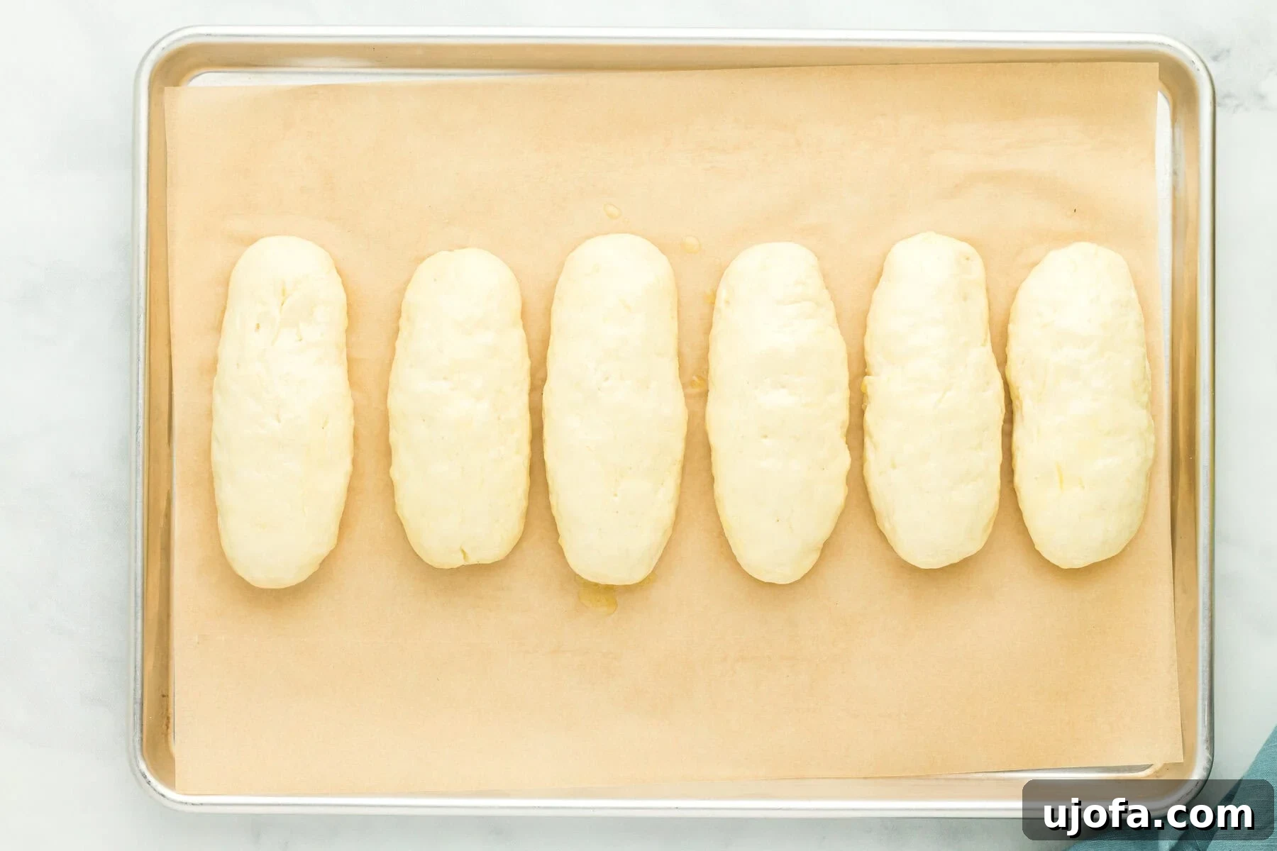 Gluten-free hot dog bun dough shaped into logs after proofing, looking noticeably larger on a parchment-lined baking sheet.