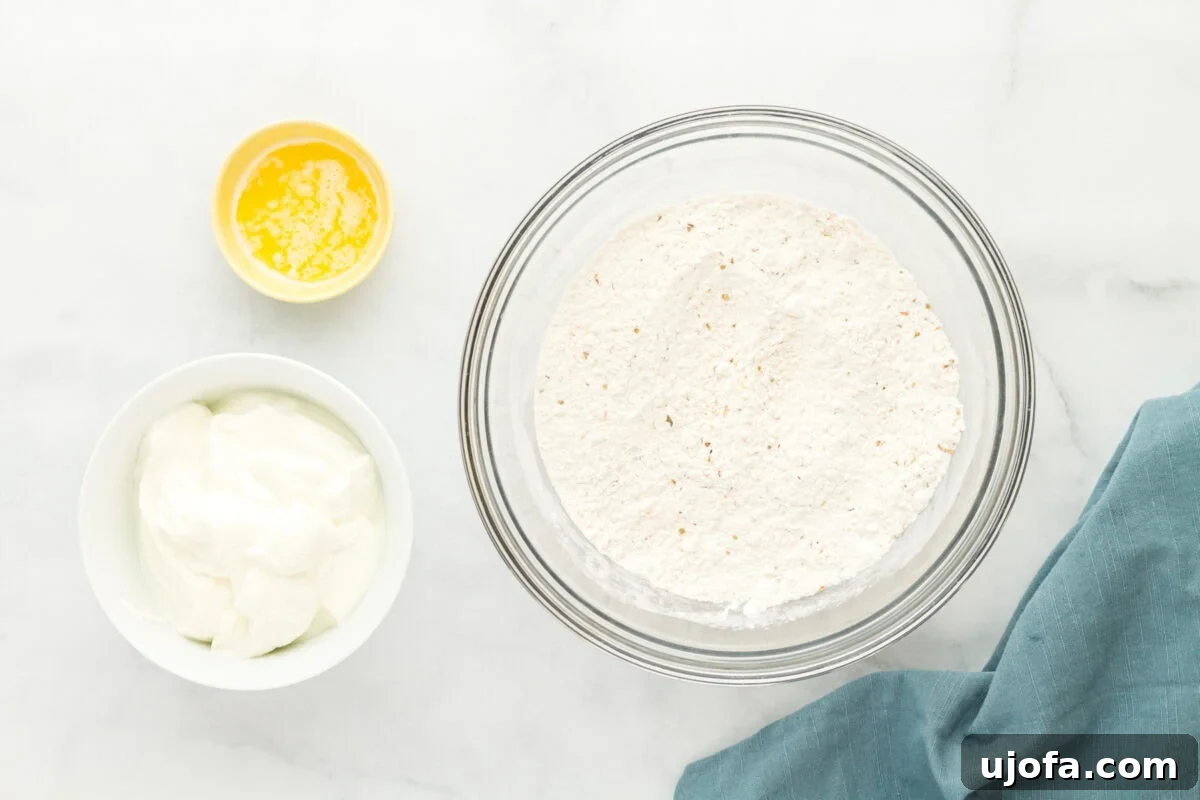 Effortless Gluten-Free Flatbread 7 Ingredients for making gluten-free flatbread, including a bowl of flour mixture, a small bowl of melted butter, and a small bowl of yogurt, arranged on a white marble surface with a blue cloth napkin