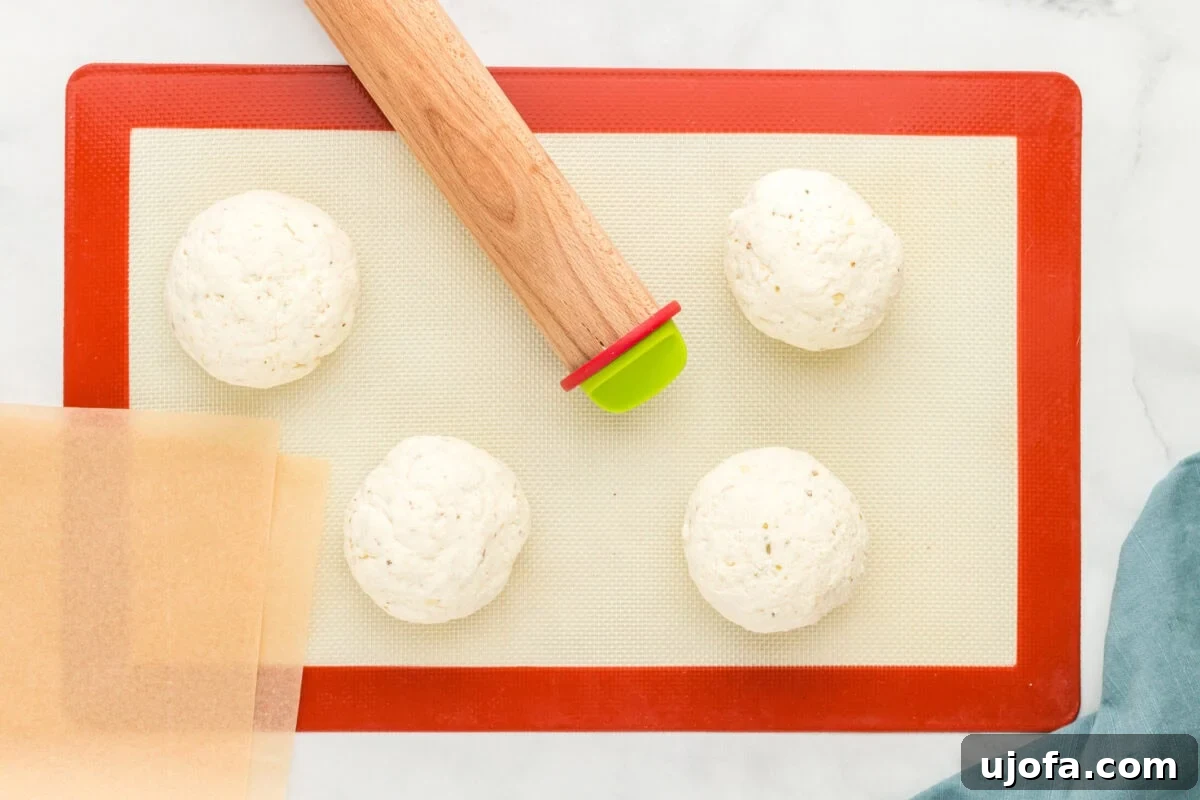 Effortless Gluten-Free Flatbread 9 Four evenly shaped gluten-free flatbread dough balls placed on a red-edged silicone baking mat with sheets of parchment paper and a wooden rolling pin nearby, on a white marble surface with a blue cloth napkin.
