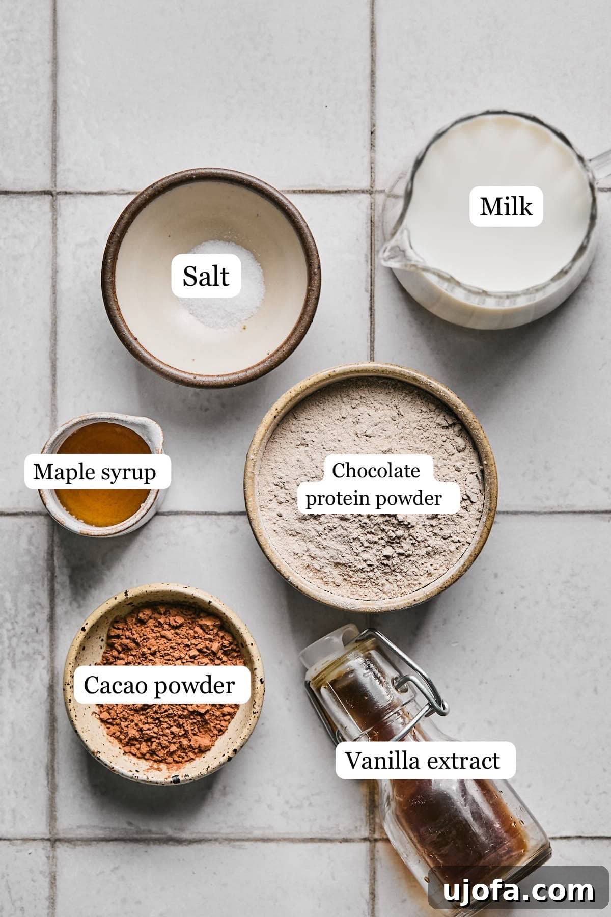 Cozy Protein-Packed Cocoa 3 Various ingredients for protein hot chocolate, including milk, protein powder, cacao, maple syrup, and vanilla extract, neatly arranged in small bowls on a wooden surface.