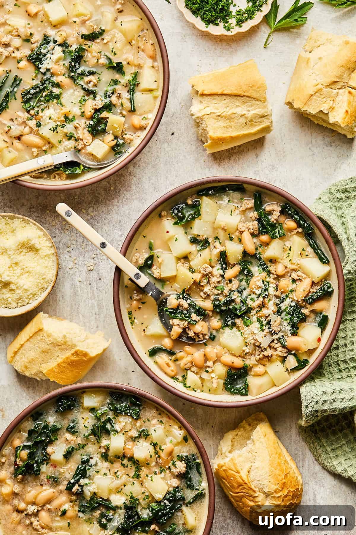 Three bowls of hearty sausage kale soup, garnished and ready to eat, surrounded by slices of crispy baguette.