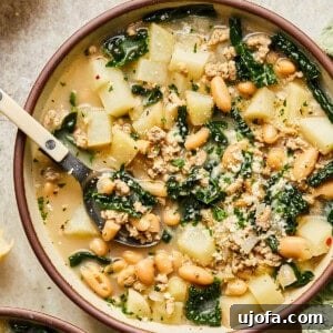 A comforting bowl of sausage, kale, and white bean soup with a spoon, surrounded by slices of crusty baguette.