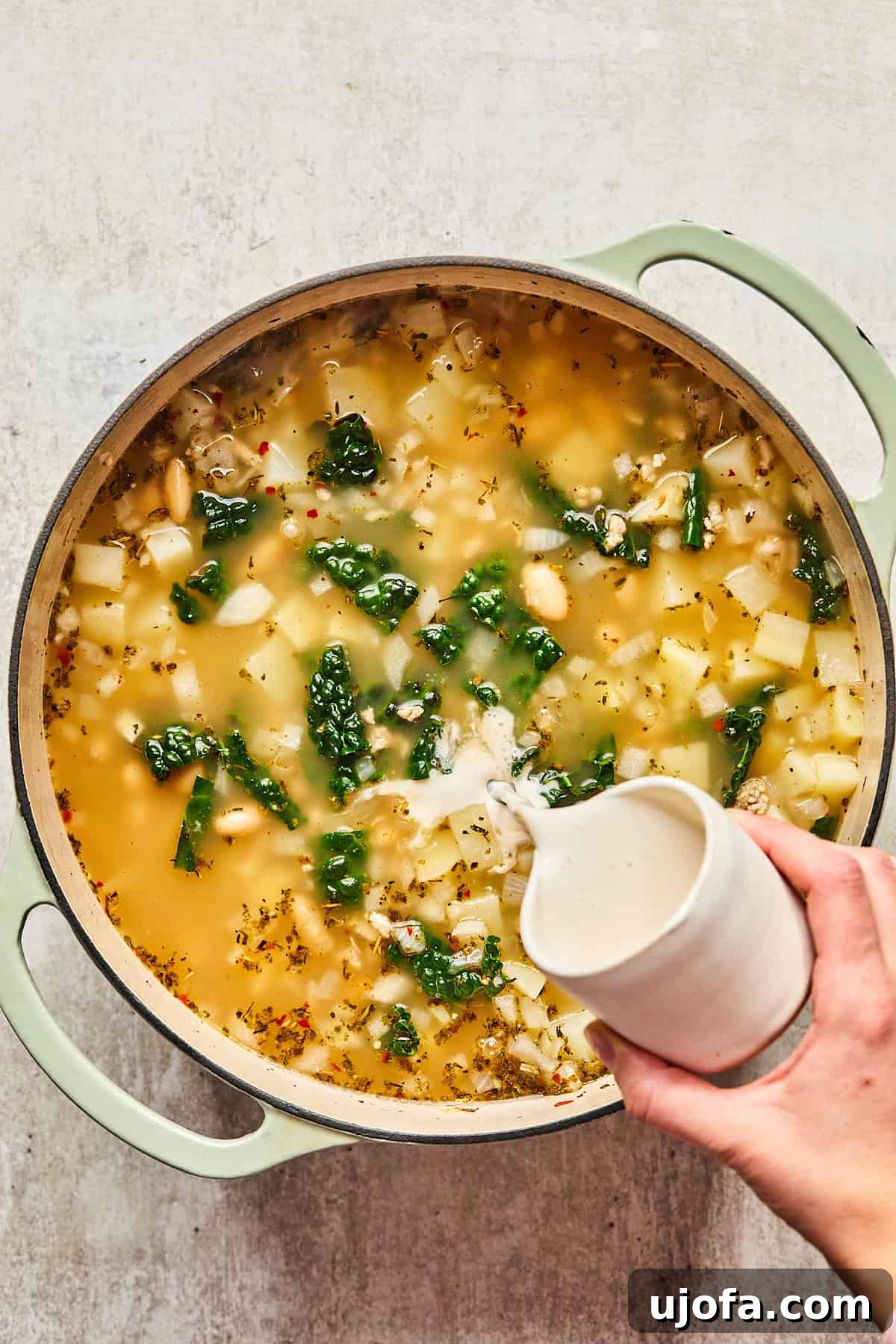 A hand pouring half & half into the finished sausage kale and white bean soup, adding a creamy touch.