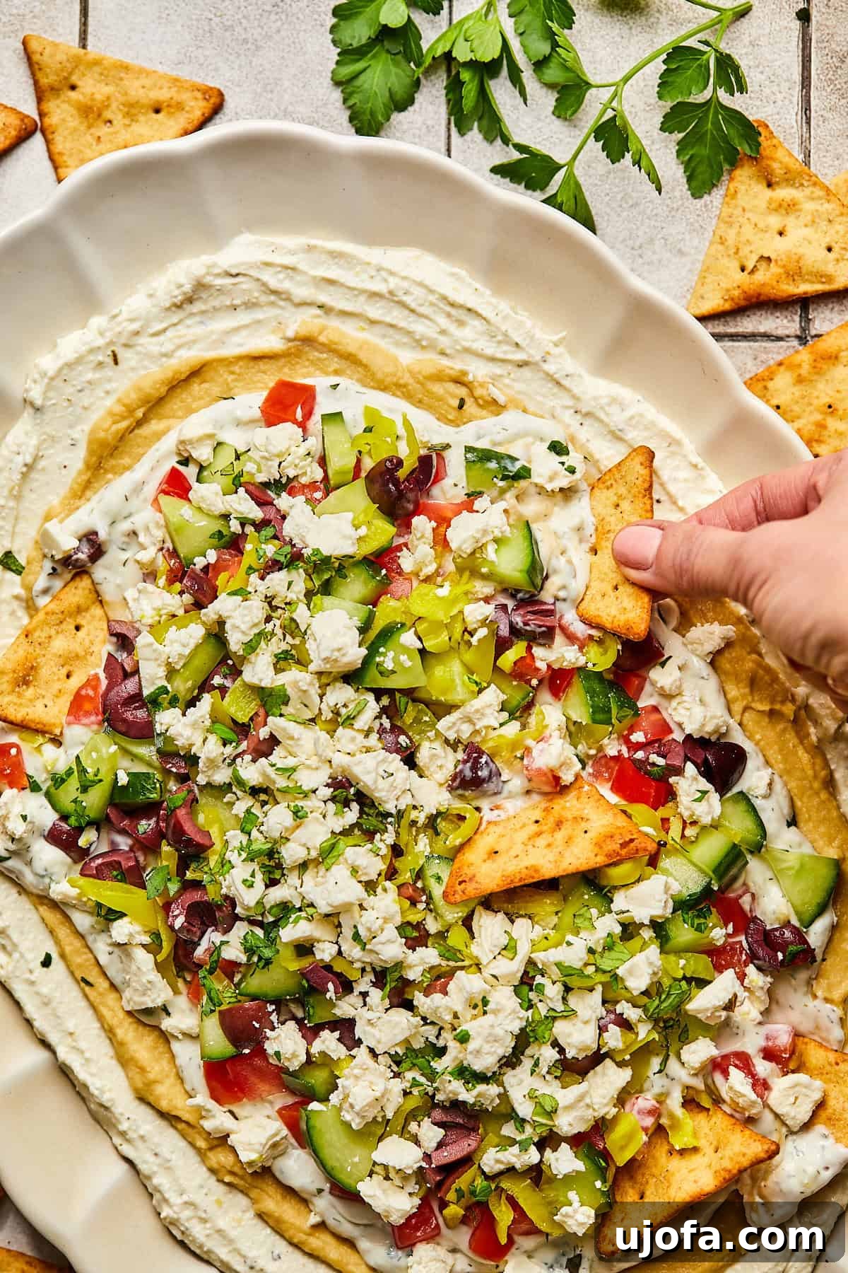 A hand reaches into a 7-layer Greek dip with a triangle pita chip, highlighting the luscious layers and inviting texture.