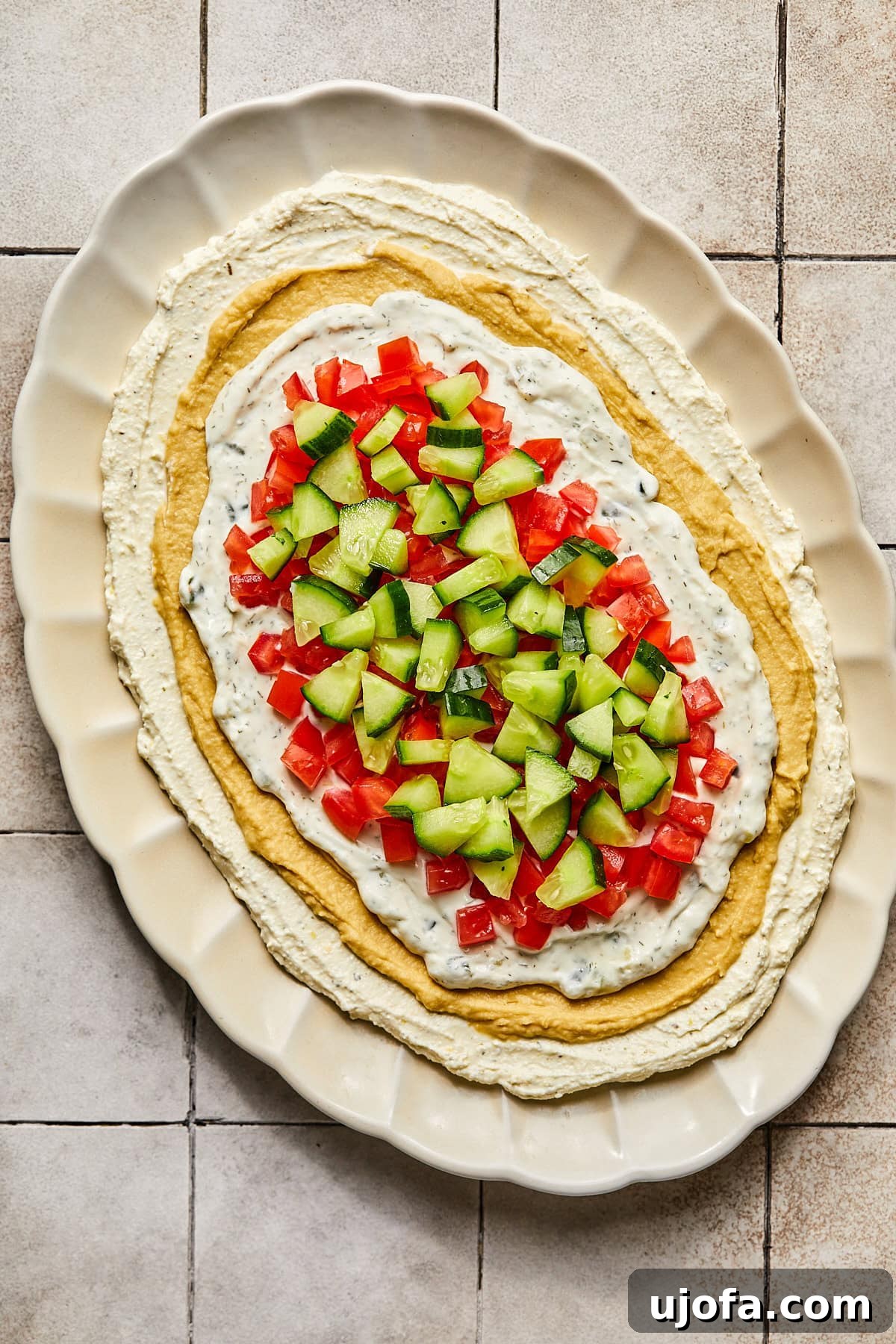 Layers of whipped feta, hummus, and tzatziki now covered with freshly diced tomatoes and cucumbers, adding color and freshness.