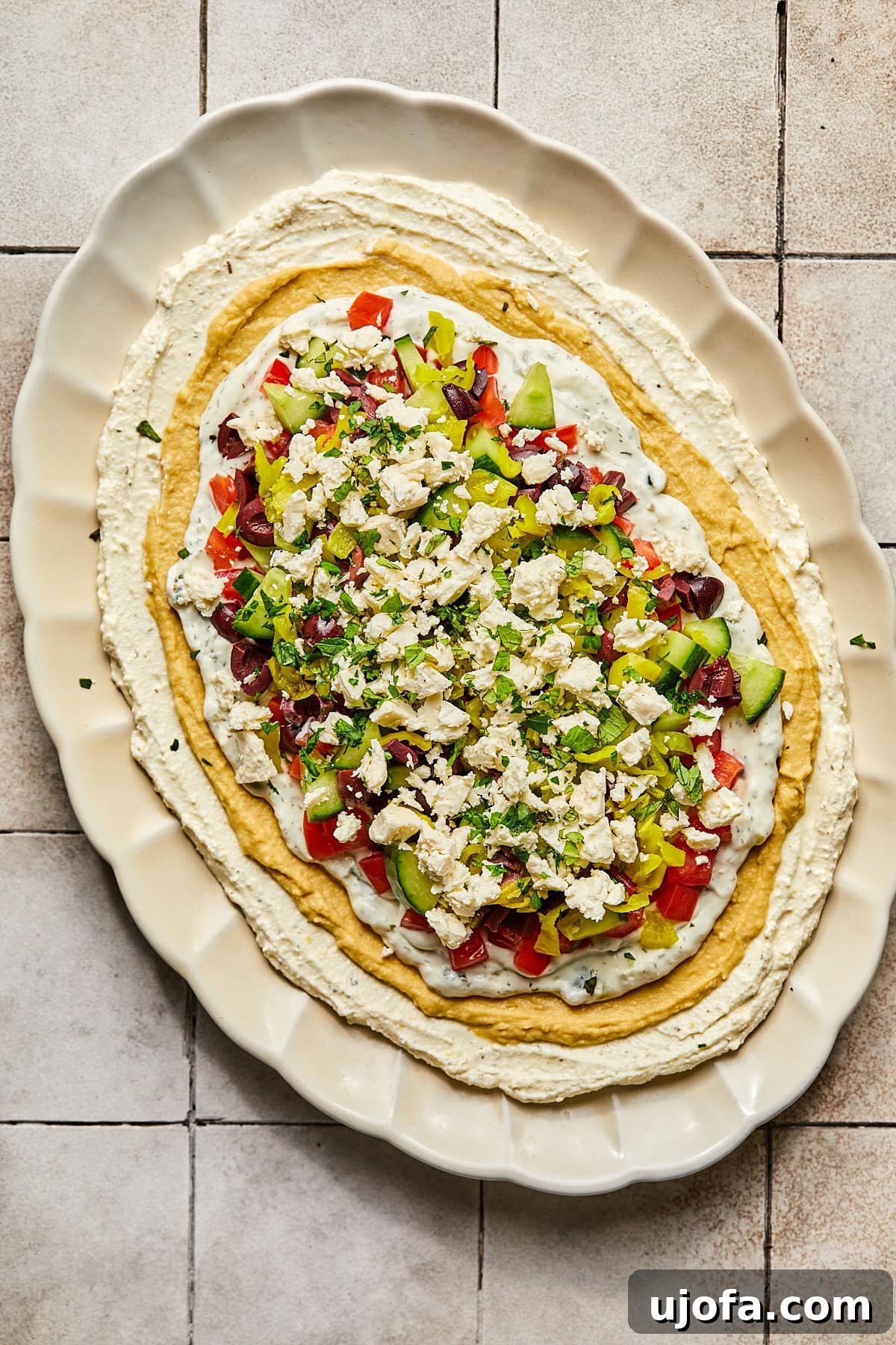 The complete Greek dip, fully layered with whipped feta, hummus, tzatziki, tomatoes, cucumbers, olives, and pepperoncinis, garnished with fresh herbs.