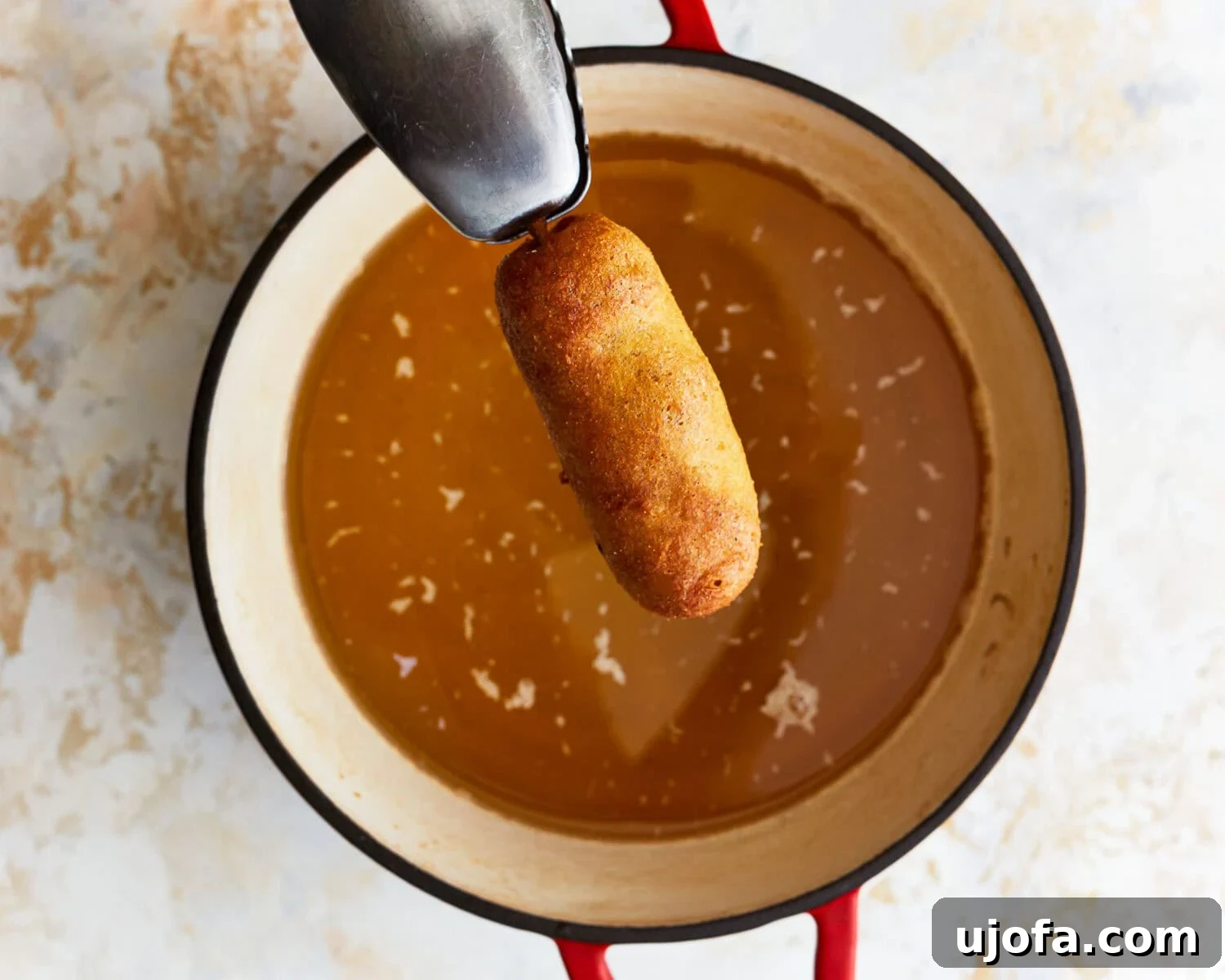 Irresistible Gluten-Free Corn Dogs 10 A cooked, golden-brown corn dog being held with tongs over a pot of hot oil, ready to be placed on paper towels.