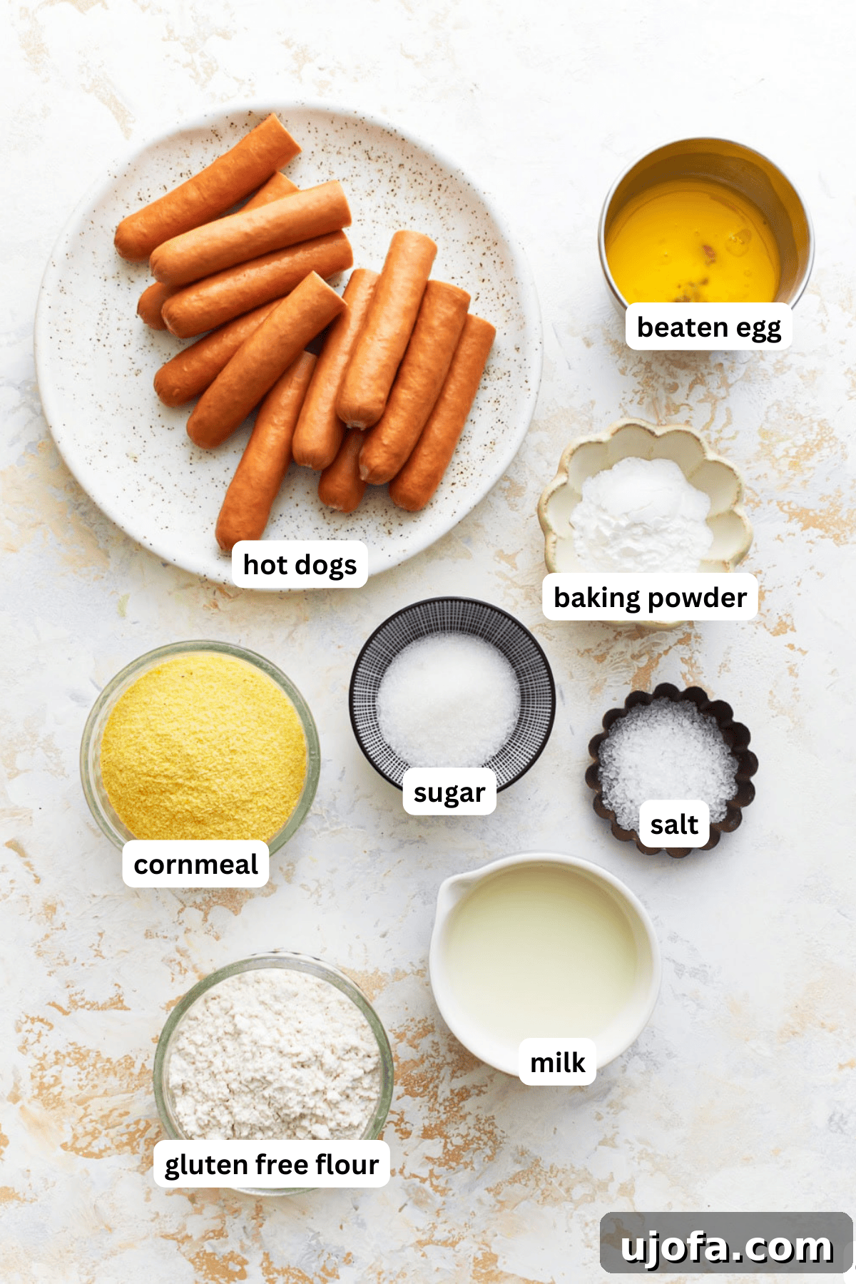 Irresistible Gluten-Free Corn Dogs 3 A flat lay photograph showing all the ingredients neatly arranged for gluten-free corn dogs: hot dogs, corn flour, GF all-purpose flour, baking powder, sugar, salt, egg, milk, and a bottle of canola oil.