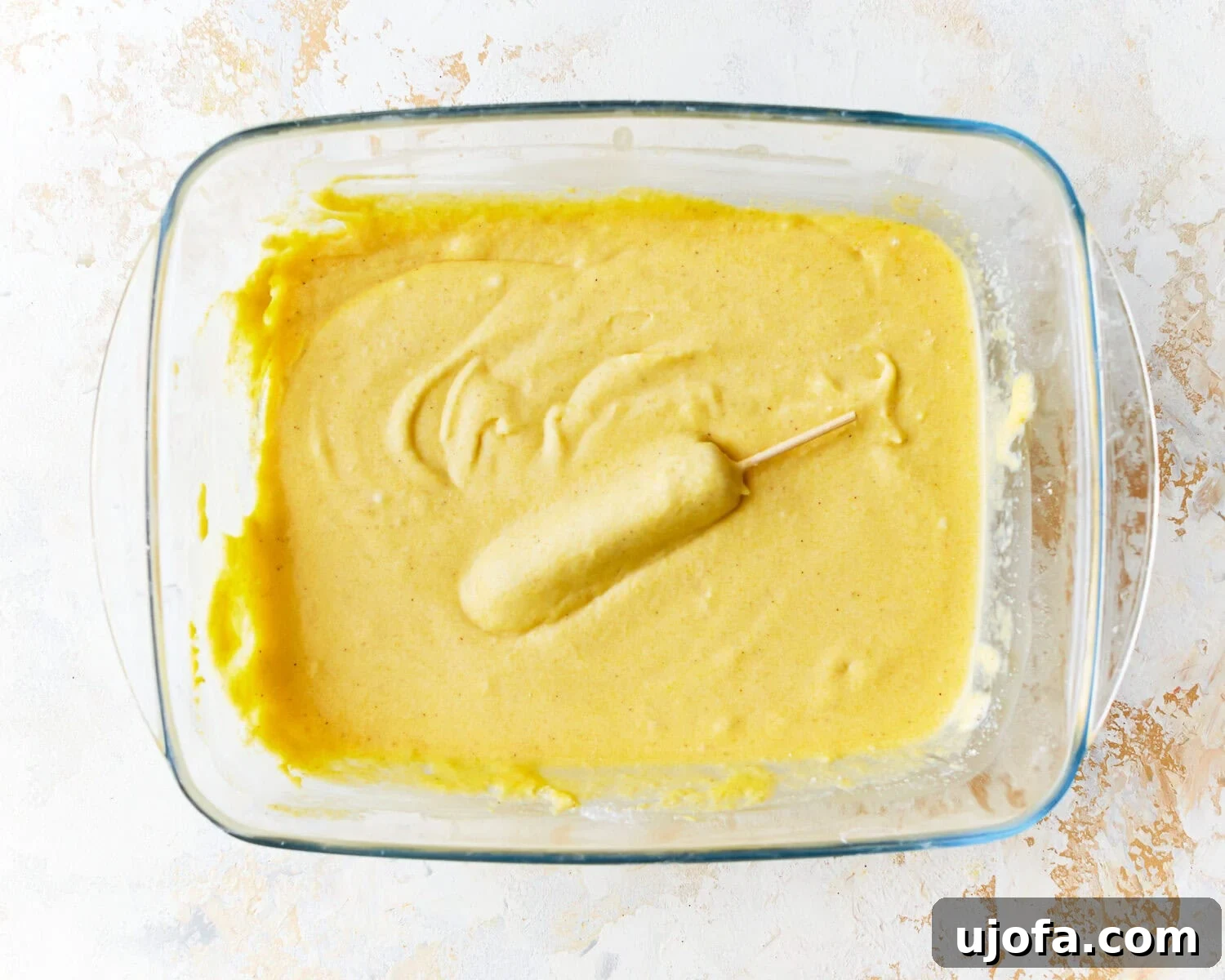 Irresistible Gluten-Free Corn Dogs 9 A hot dog on a skewer being dipped into the corn dog batter in a glass dish, ensuring full coating.