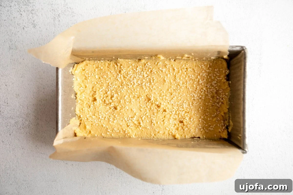 Raw coconut flour bread dough, spread evenly in a parchment-lined loaf pan, topped with sesame seeds, ready for baking.
