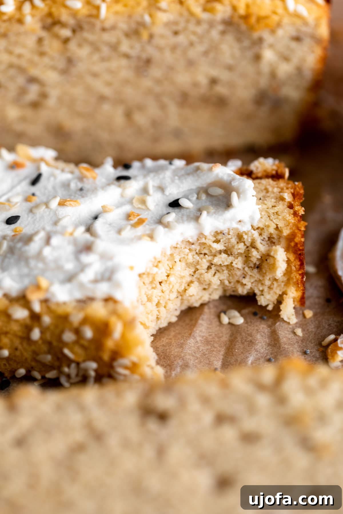 A slice of coconut flour bread topped with cream cheese and everything bagel seasoning, with a bite taken out, resting on a cutting board.