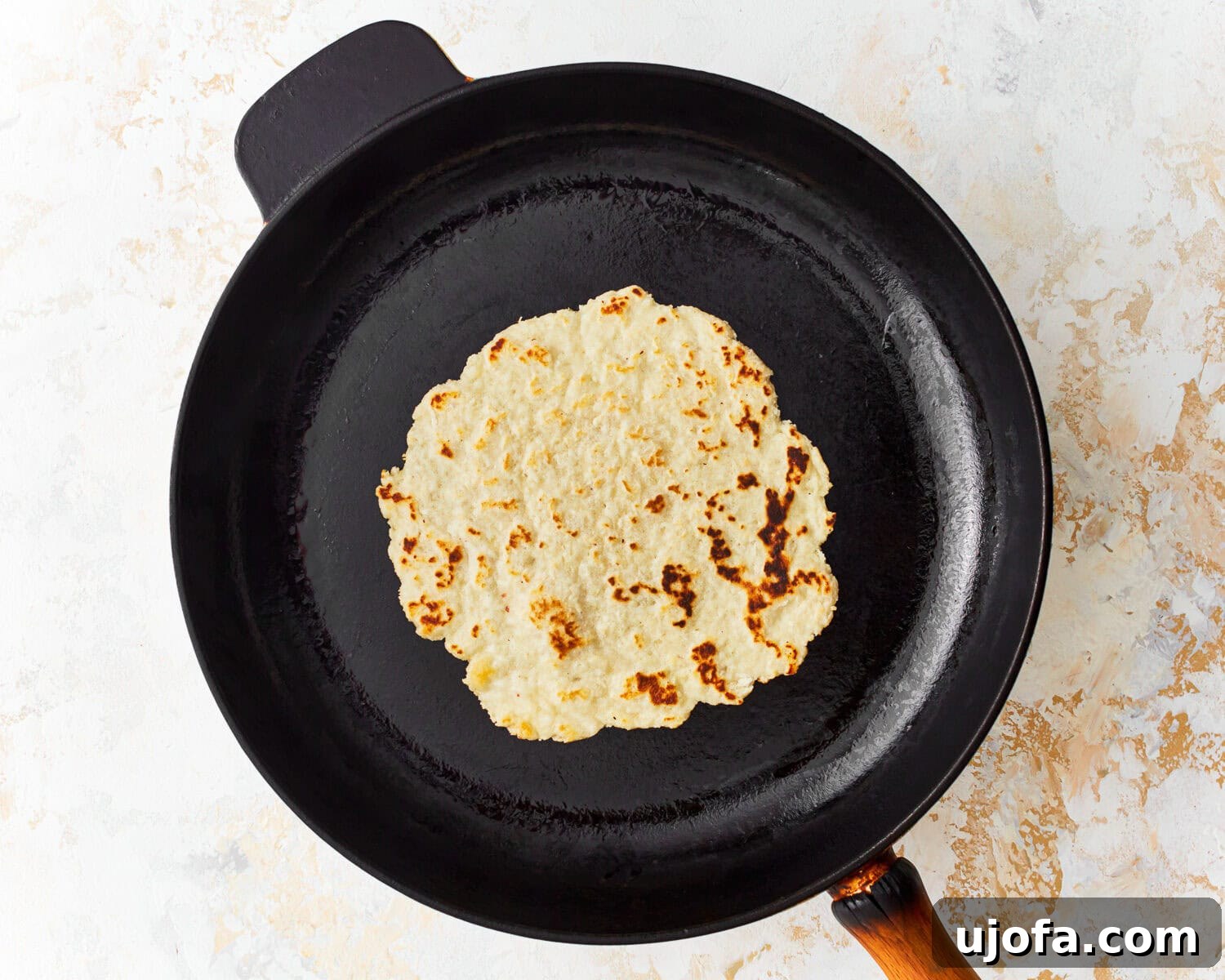 Overhead view of an almond flour tortilla being flipped in a skillet, revealing golden-brown spots.