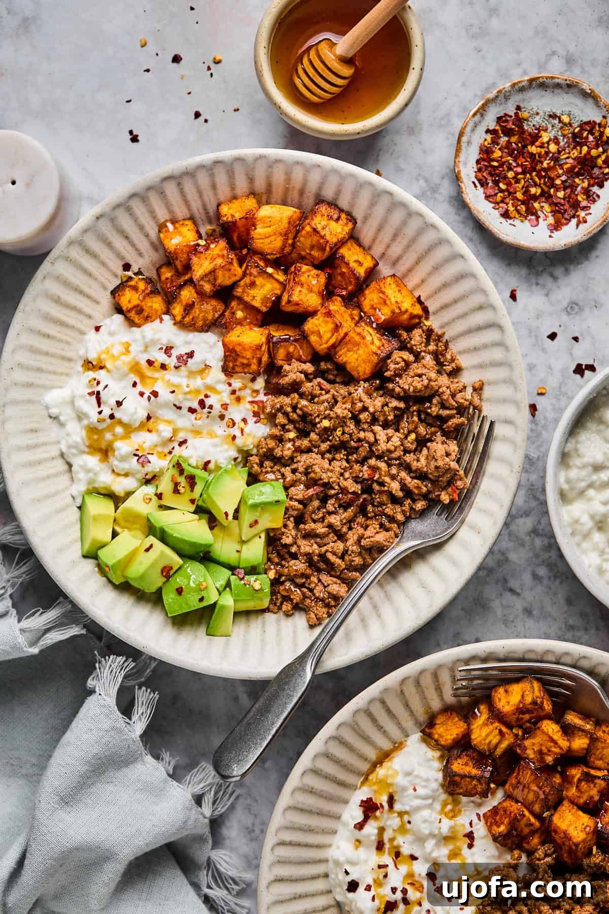 A gourmet Cottage Cheese Taco Bowl expertly arranged with seasoned ground meat, tender roasted sweet potatoes, fresh avocado slices, and creamy cottage cheese, elegantly finished with a scattering of red pepper flakes. A fork rests casually beside the bowl, with complementary items like hot honey, extra red pepper flakes, a napkin, and a small decorative candle completing the inviting tableau.
