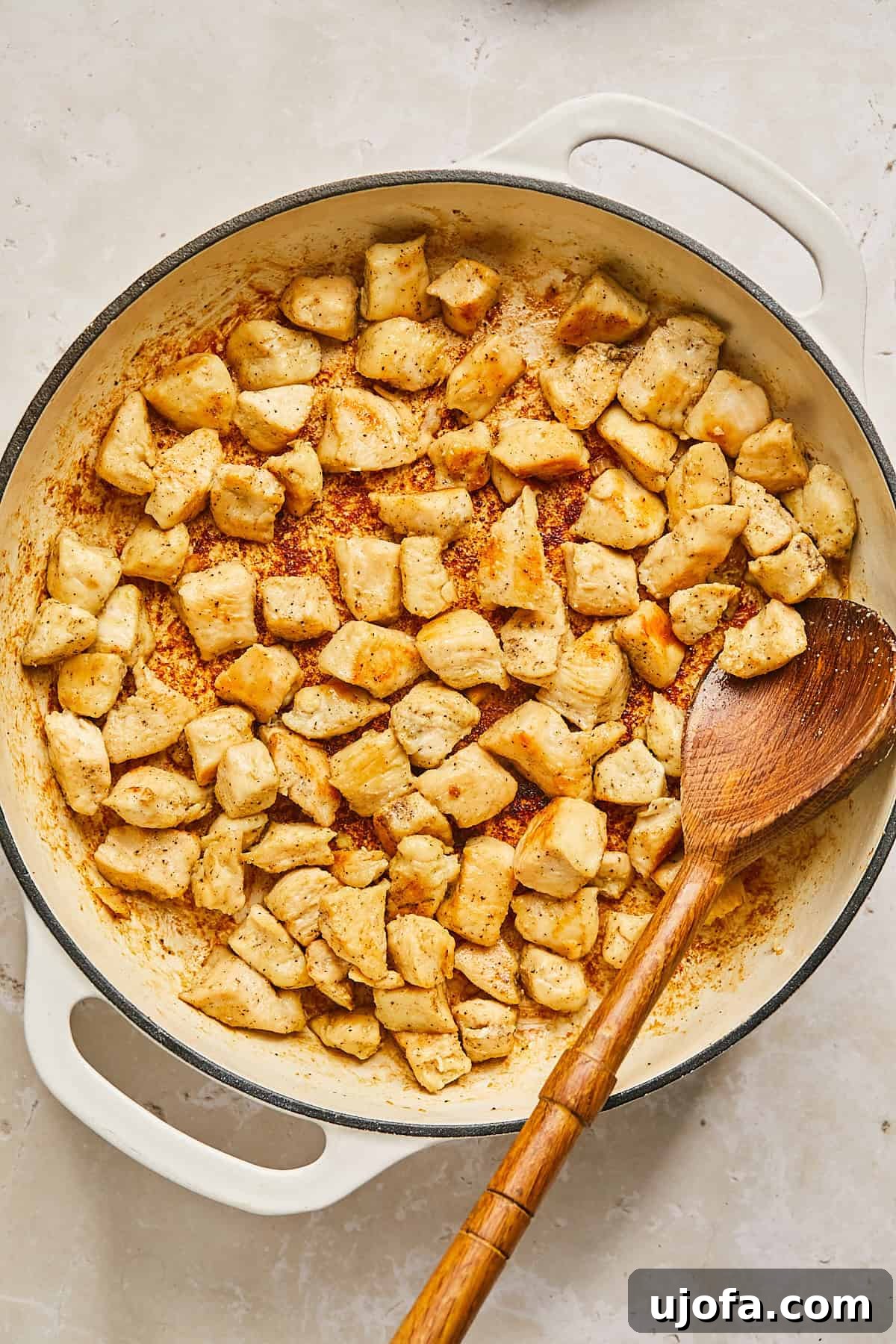A white skillet filled with cooked, golden-brown chicken pieces, seasoned with spices. A wooden spoon rests in the skillet, suggesting the chicken has been stirred or sautéed. The skillet is on a neutral-colored countertop.