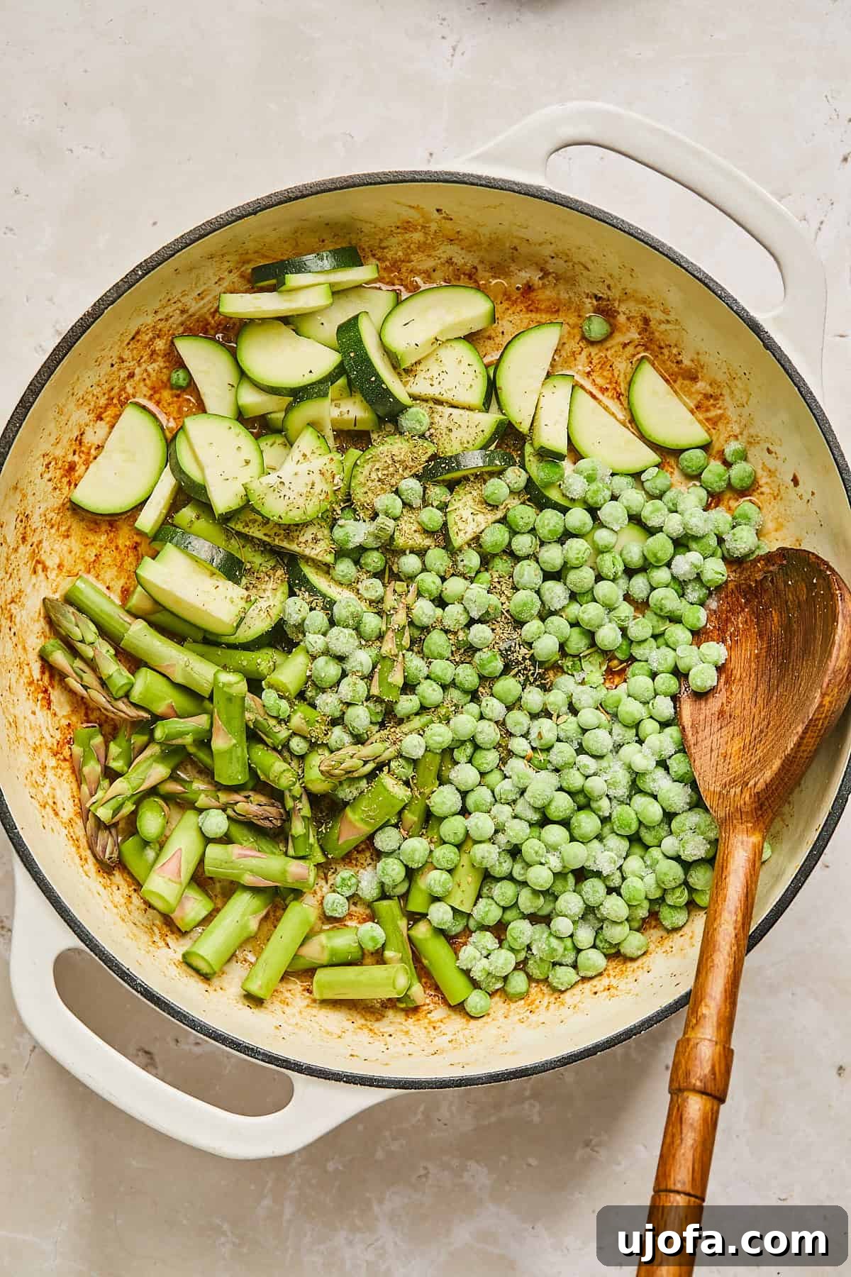 A white pot filled with chopped asparagus, zucchini slices, and frozen peas, placed on a light beige surface. A wooden spoon rests inside the pot, ready to stir the vibrant, fresh ingredients.