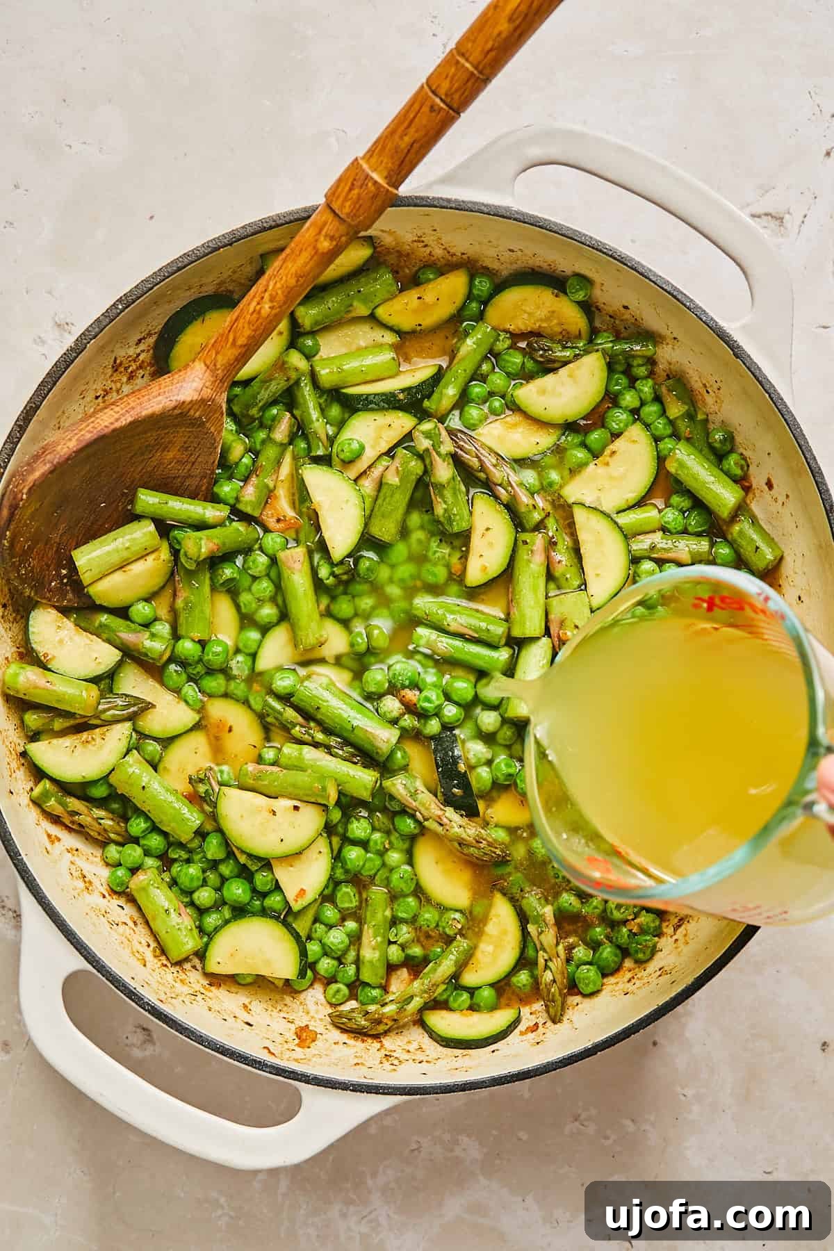 A cast iron pan filled with a mixture of sliced zucchini, asparagus, and peas is placed on a light surface. A wooden spoon rests in the pan, and a measuring cup is pouring broth into the mixture.