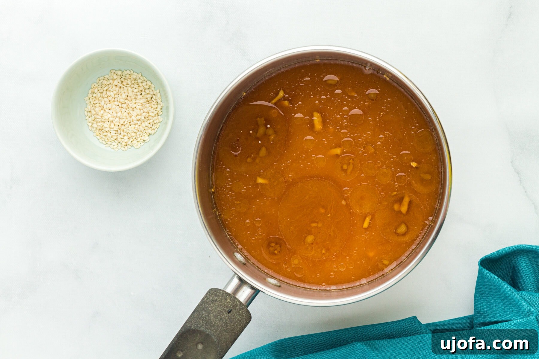 Overhead view of cornstarch slurry mixed into gluten free teriyaki sauce in a saucepan next to a bowl of sesame seeds.