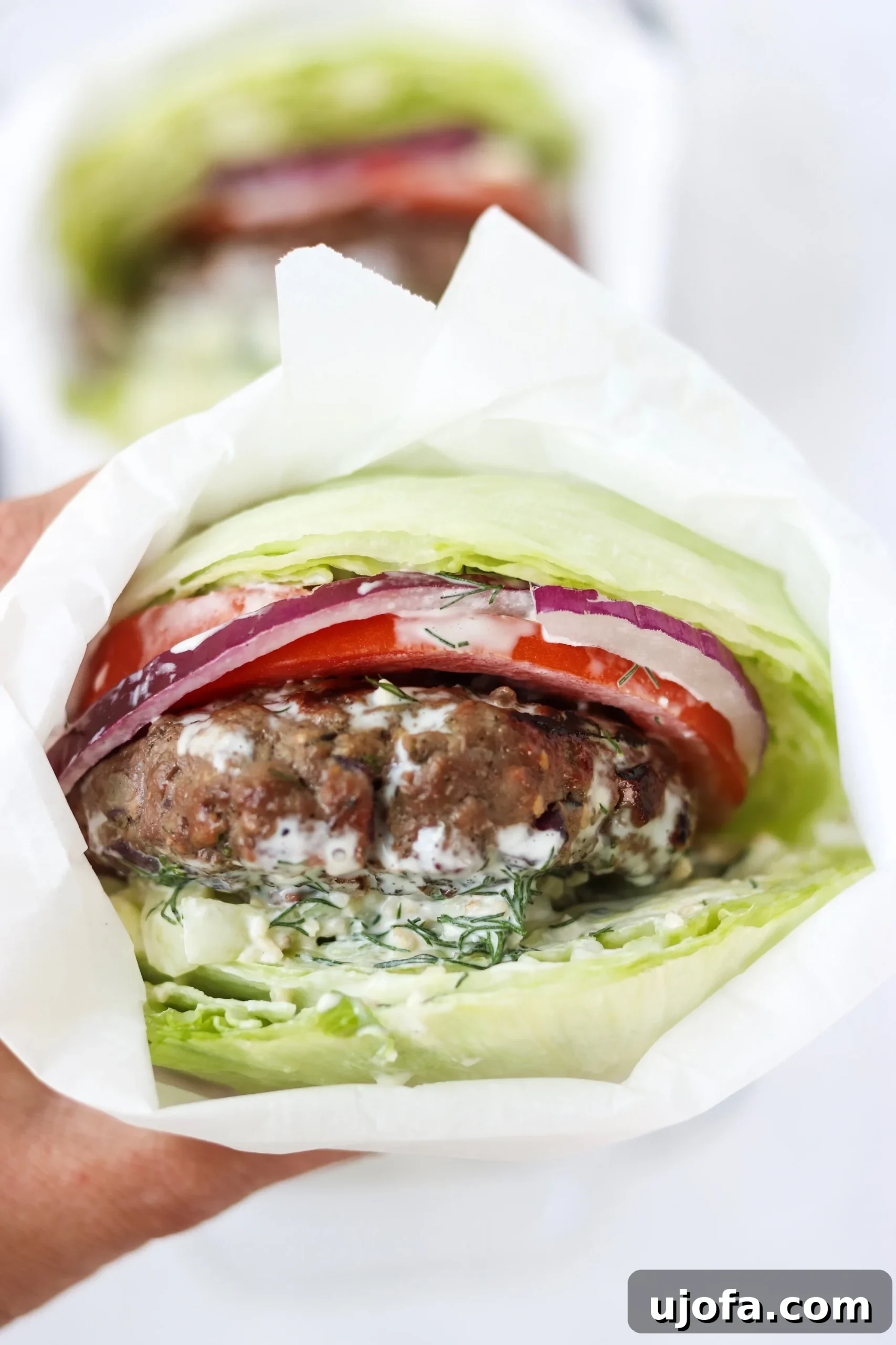 A vibrant platter of Greek burger toppings including sliced tomatoes, red onions, lettuce, and a bowl of dairy-free tzatziki sauce, alongside grilled patties.
