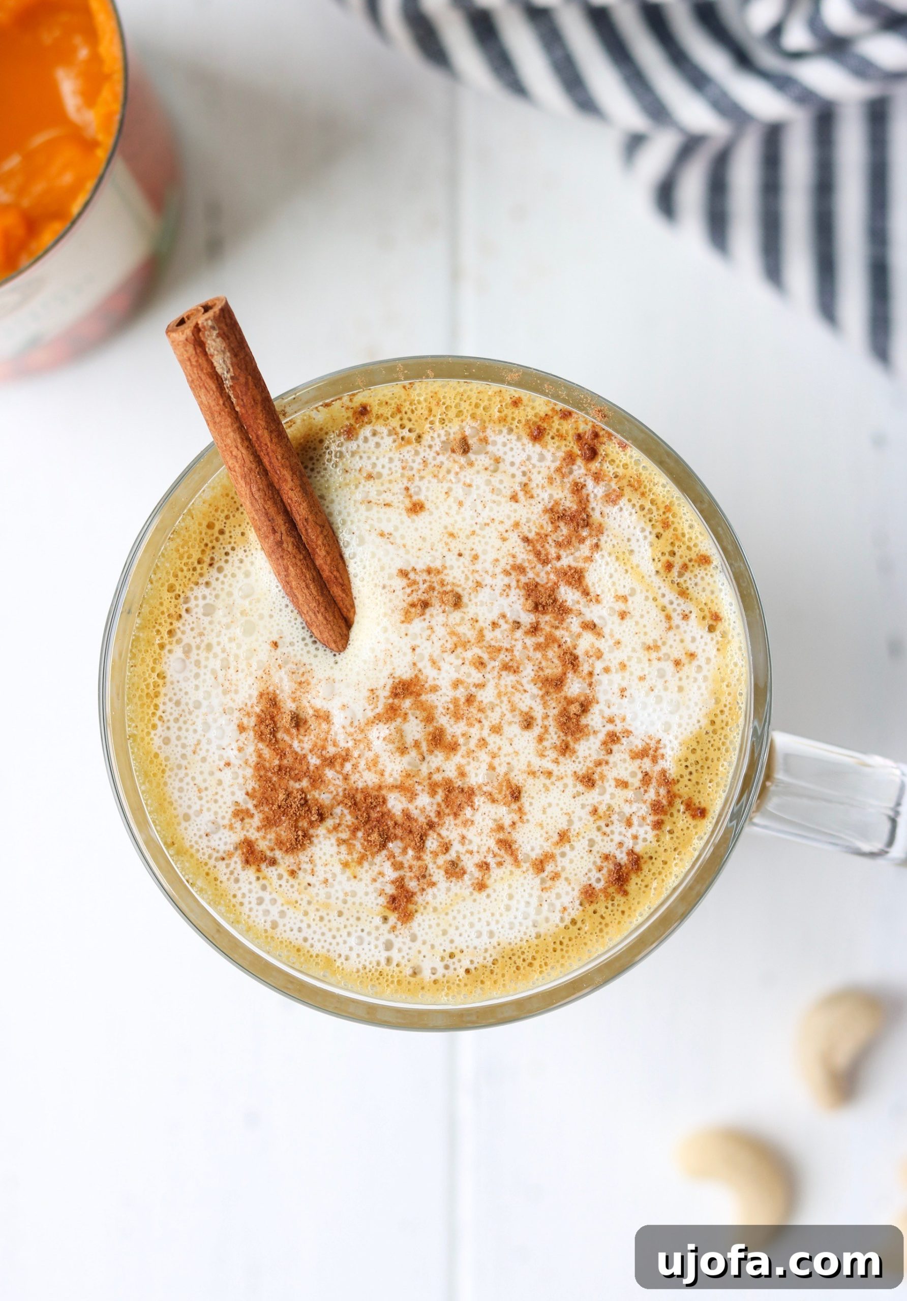 A warm mug of Creamy Cashew Pumpkin Spice Latte on a wooden surface, with a spoon and spices nearby.