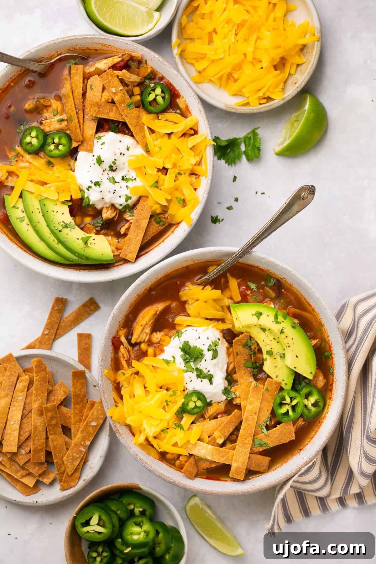 Two bowls of chicken tortilla soup on a light gray countertop garnished with shredded cheese, tortilla strip chips, avocado slices, jalapenos, and sour cream.