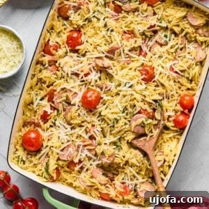 chicken sausage orzo bake in a casserole dish with a wooden spoon surrounded by tomatoes, cheese, and a pink napkin