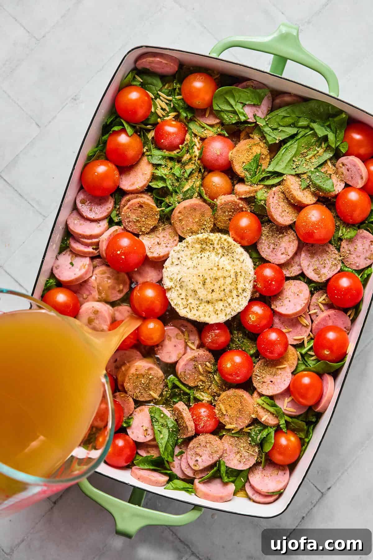 Kremamsı Boursin Orzo Tavuk Sosisli Tek Tencere Keyfi 5 Unbaked orzo, sliced chicken sausage, cherry tomatoes, and spinach arranged around the Boursin cheese in a baking dish, with chicken broth being poured over.