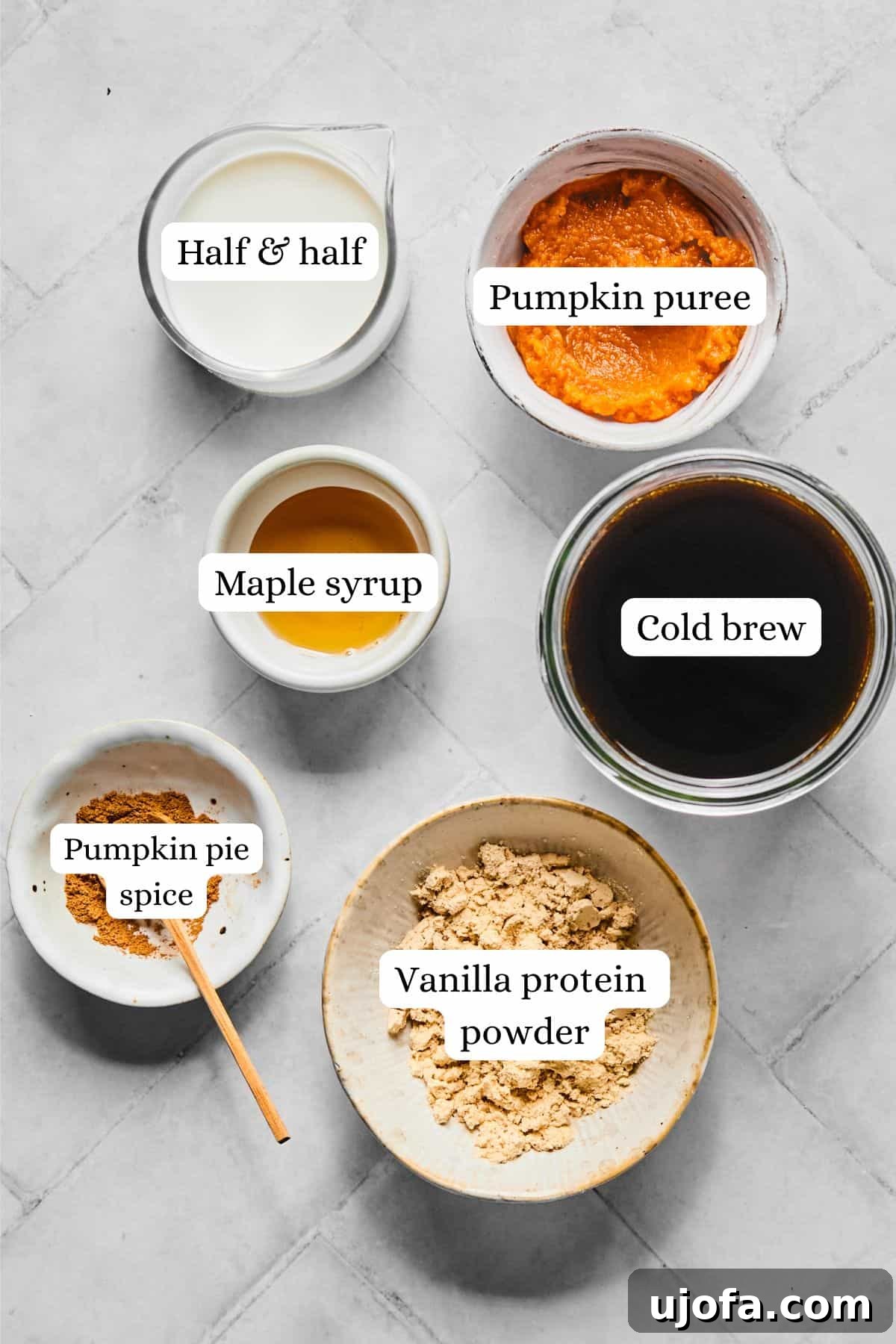 Individual ingredients for pumpkin cold foam, including pumpkin puree, vanilla protein powder, maple syrup, half and half, pumpkin pie spice, and cold brew coffee, neatly arranged in small nesting bowls.