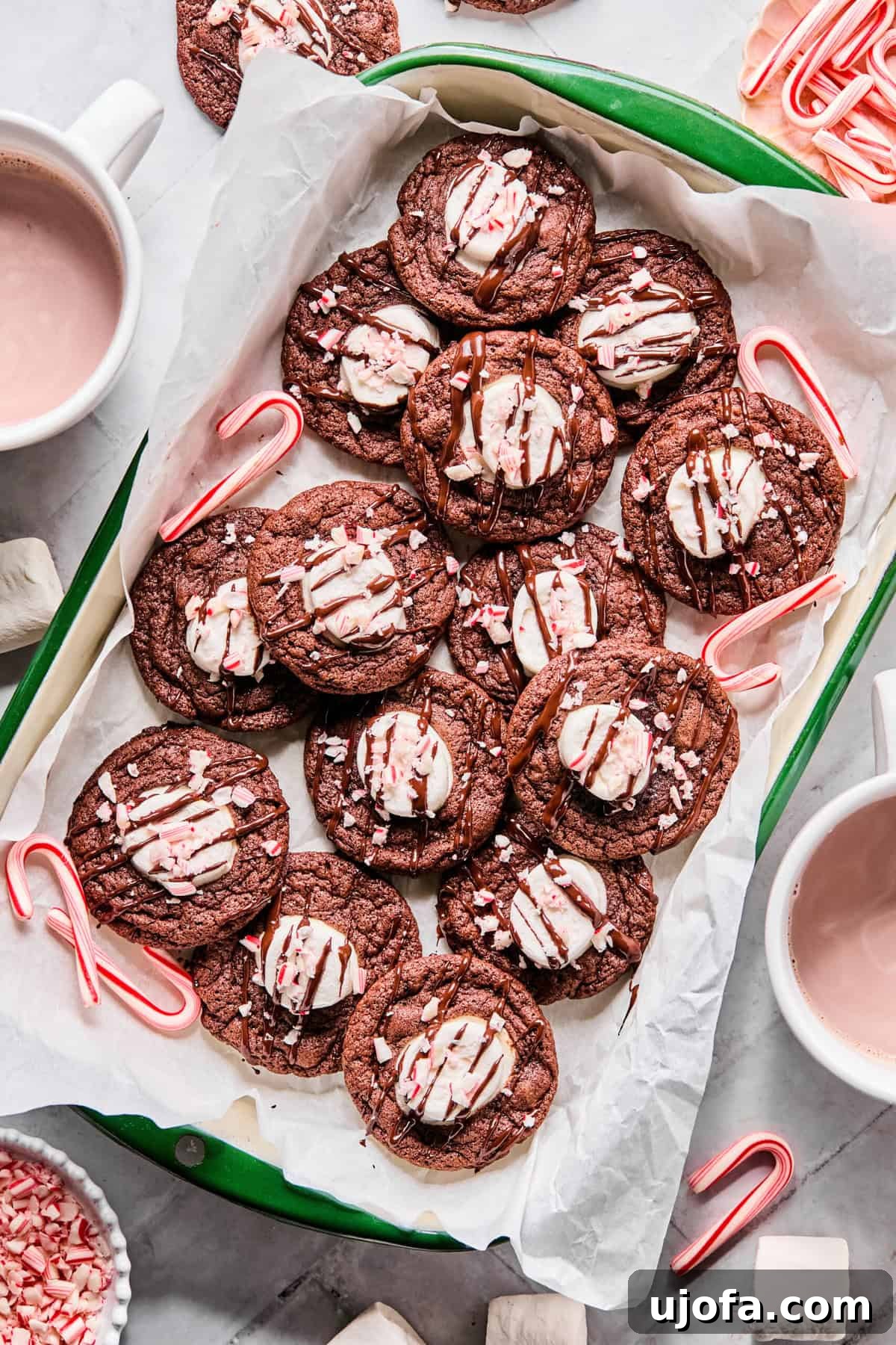 Effortless Gluten-Free Hot Chocolate Cookies 8 A festive arrangement of gluten-free hot cocoa cookies on a green parchment-lined tray, surrounded by mini candy canes, with a steaming mug of hot chocolate in the background.