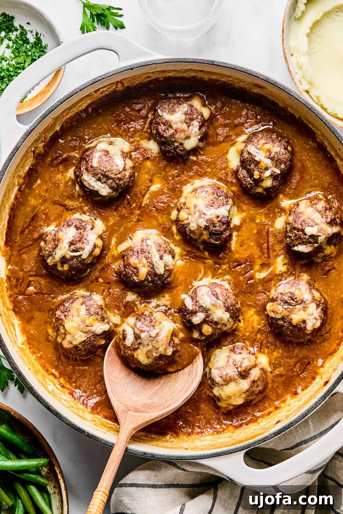 Savory French Onion Meatballs with Rich Gravy 11 Baked French onion meatballs in a large oven-safe skillet, with melted, golden-brown Gruyère cheese. A wooden spoon is scooping out a meatball.