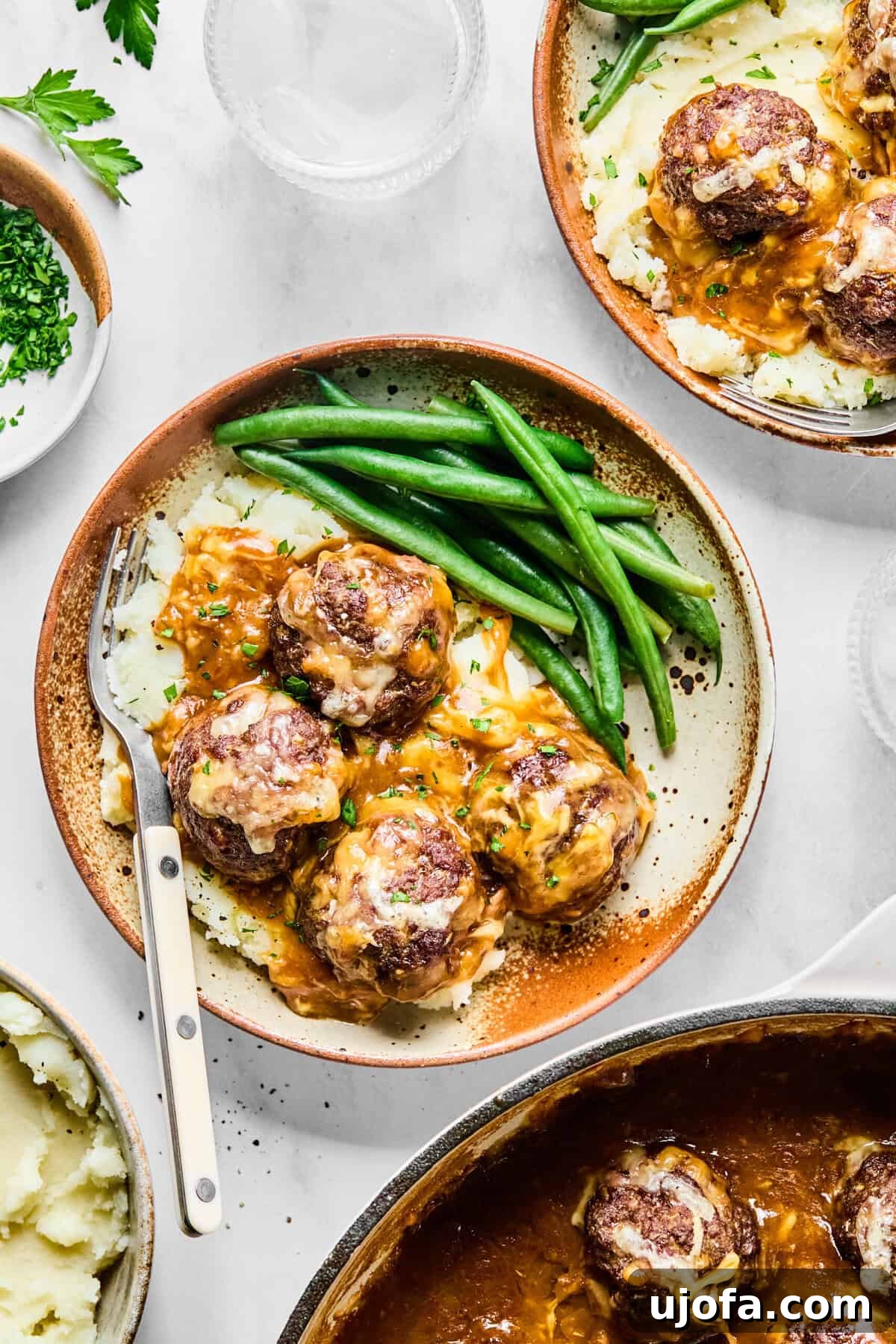 Savory French Onion Meatballs with Rich Gravy 12 Plated French onion meatballs with onion gravy, served elegantly over a bed of mashed potatoes and fresh green beans, on a light gray background.