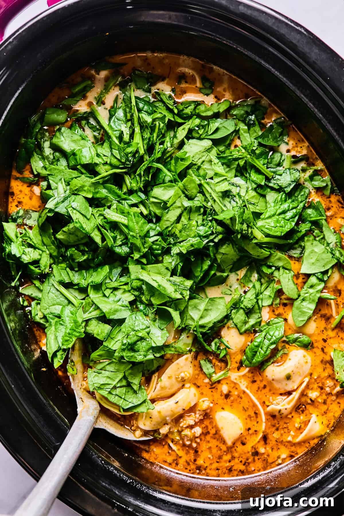 Fresh baby spinach being stirred into the hot, creamy sausage tortellini soup in the slow cooker.