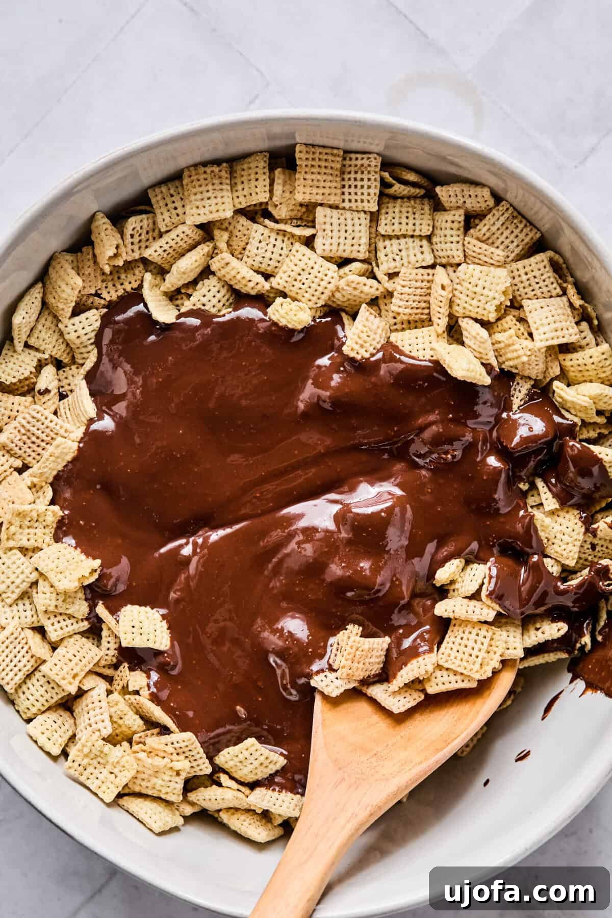 A large mixing bowl filled with Rice Chex cereal topped with melted chocolate and peanut butter, with a wooden spoon resting inside ready to mix.